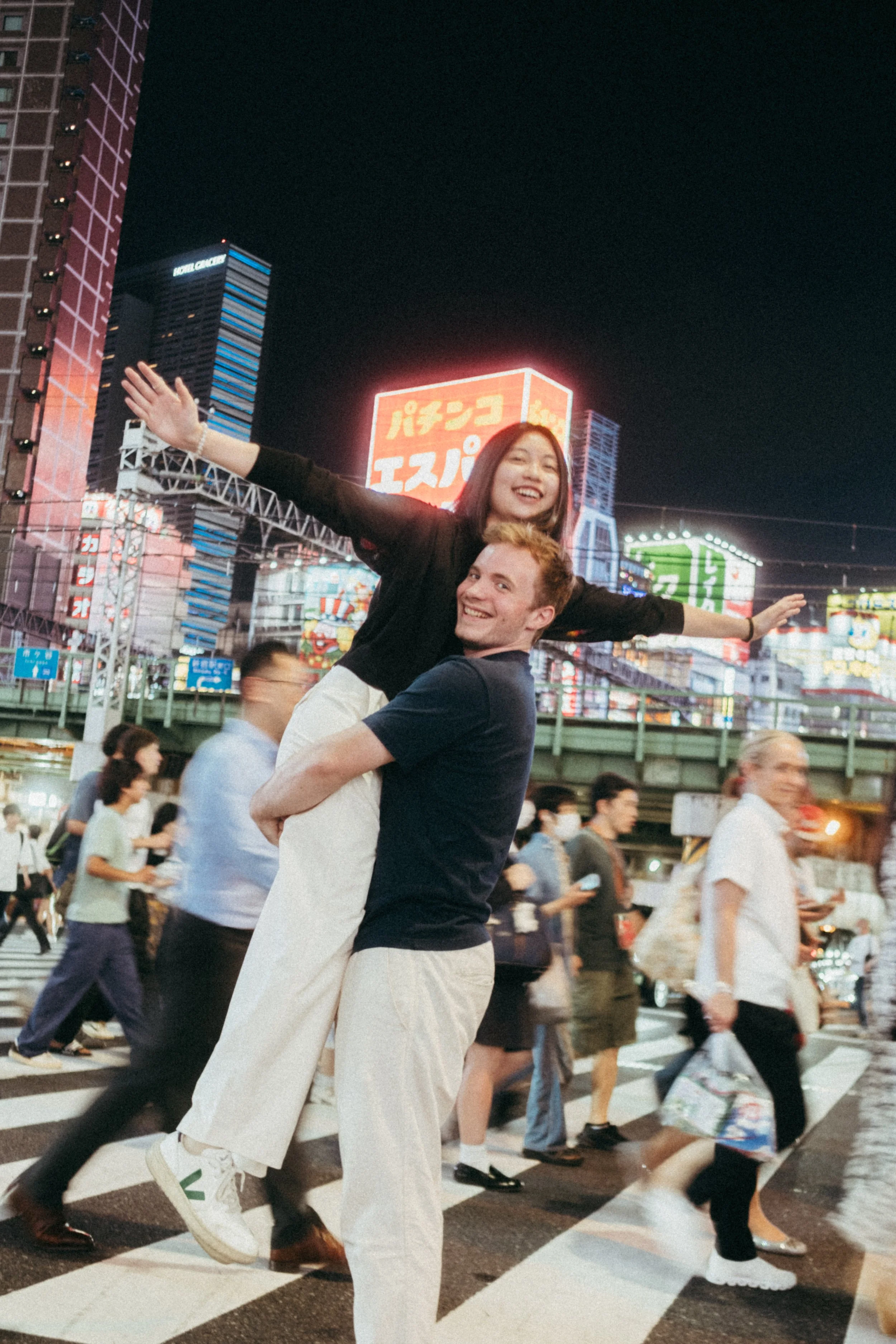 A young man is carrying a young woman on his shoulders as they walk across a busy city street at night, with neon signs and buildings in the background.