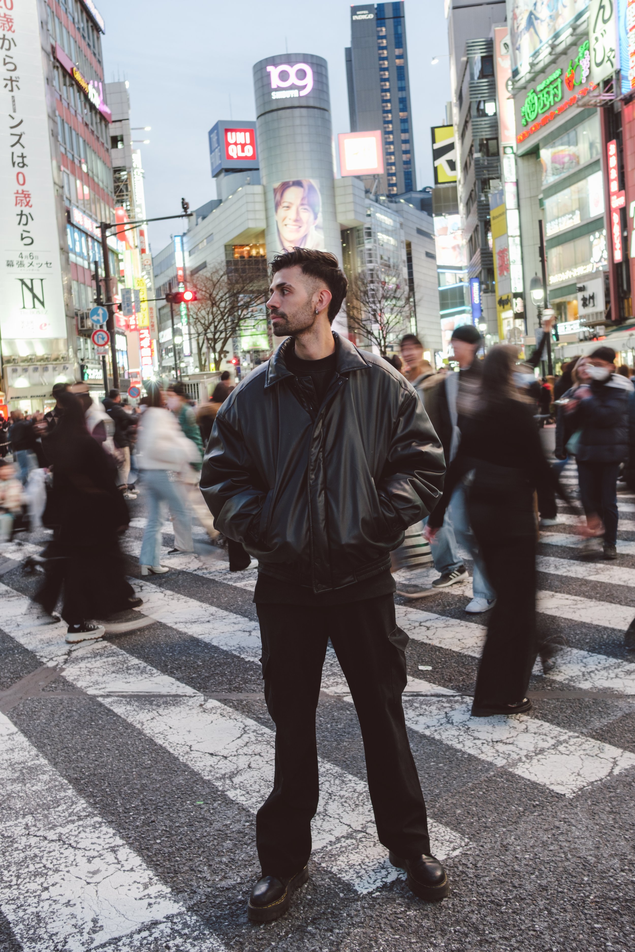 A man with dark hair and a beard wearing a black leather jacket and black pants stands in the middle of a busy city street at dusk, surrounded by blurred pedestrians and tall buildings with bright neon signs and billboards.