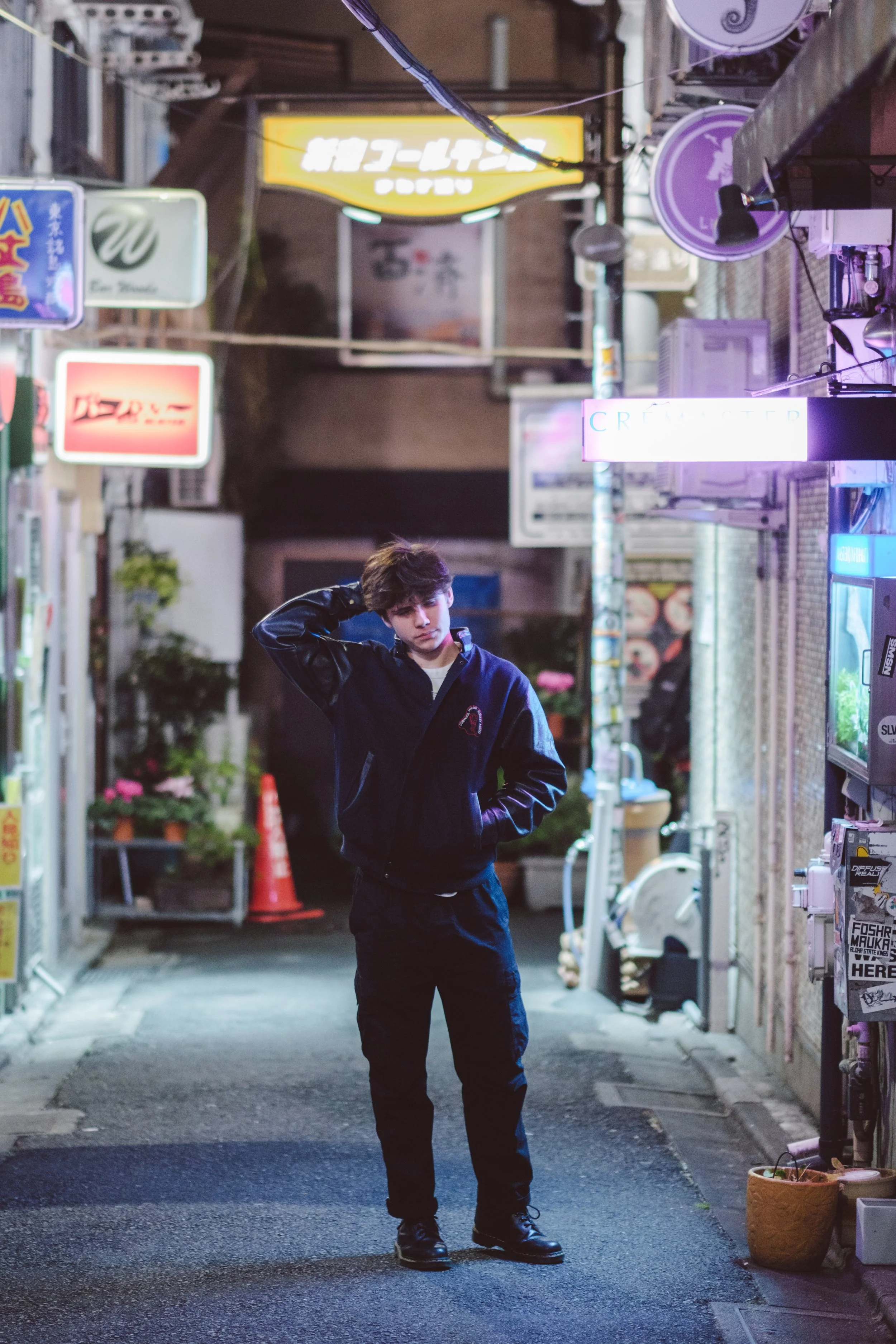 A young man with dark hair, wearing a black jacket and black pants, standing in a dimly lit alley with neon signs and storefronts. He is looking slightly downward with his hand behind his head. The alley has various signs with Japanese characters and