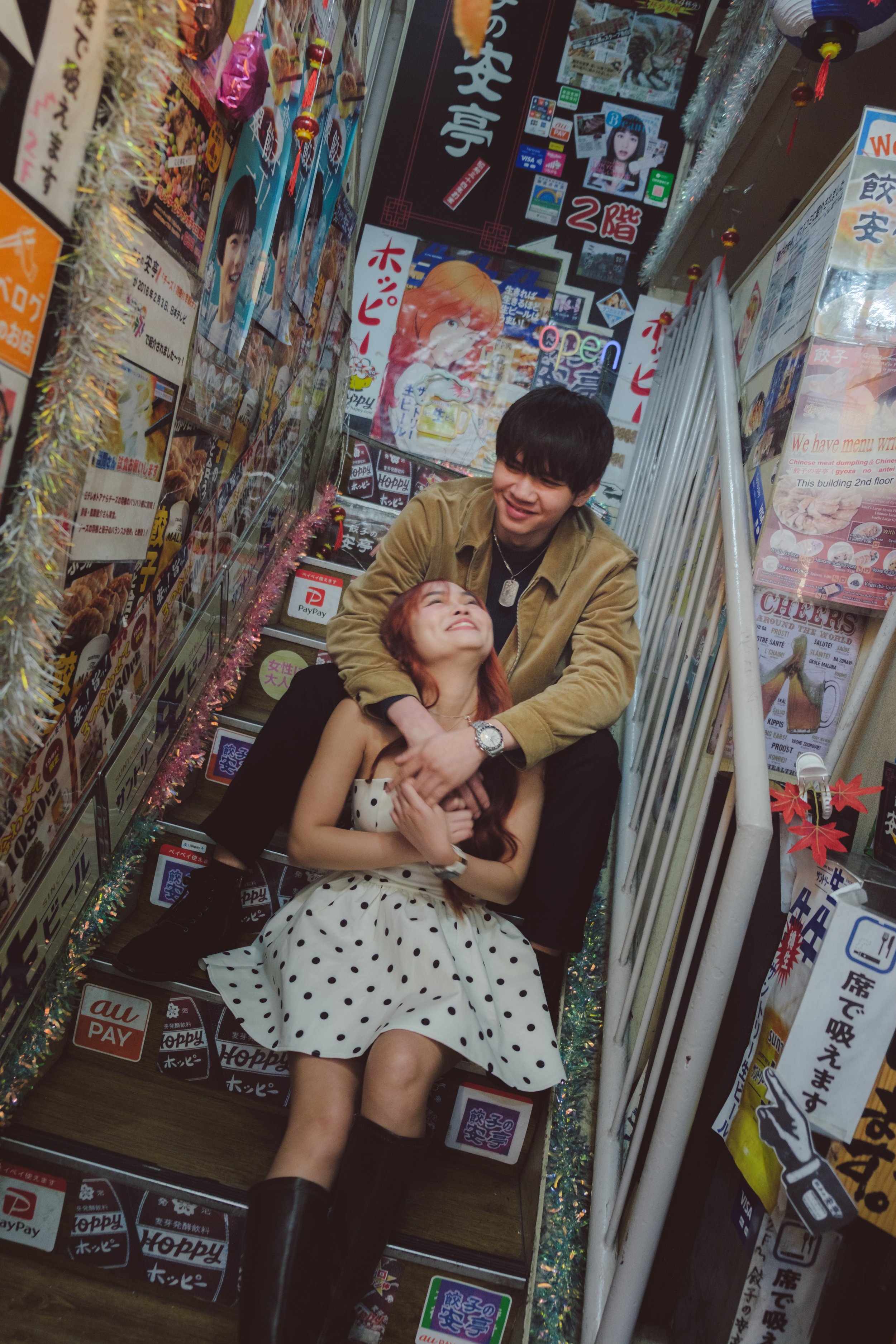 A young man and woman sitting on the floor surrounded by Japanese restaurant advertisements and colorful decorations, with the woman looking up at the man, both smiling.