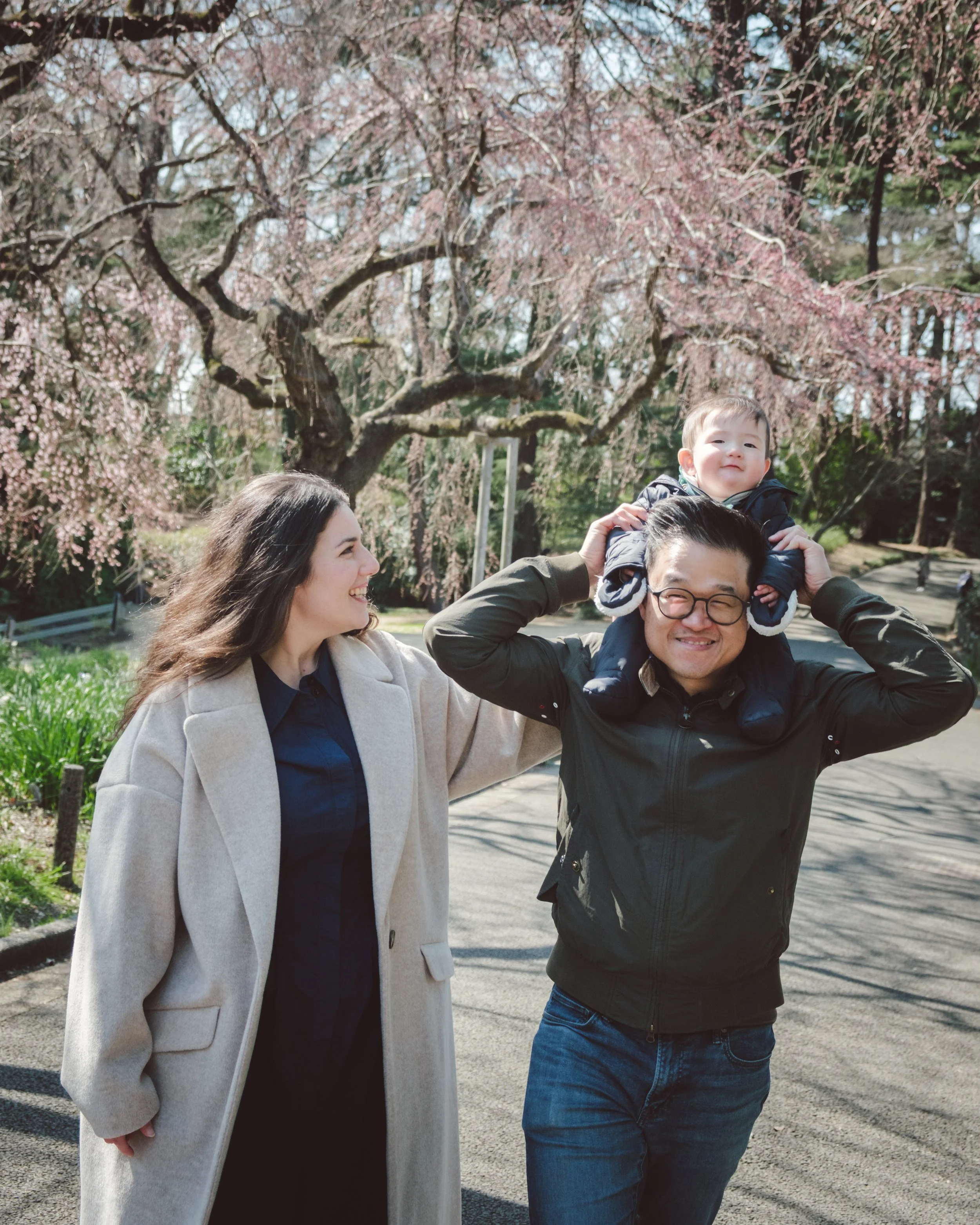 A family of three outdoors, with a young boy sitting on his father's shoulders while the mother smiles and looks at them. Cherry blossom trees are in full bloom in the background.
