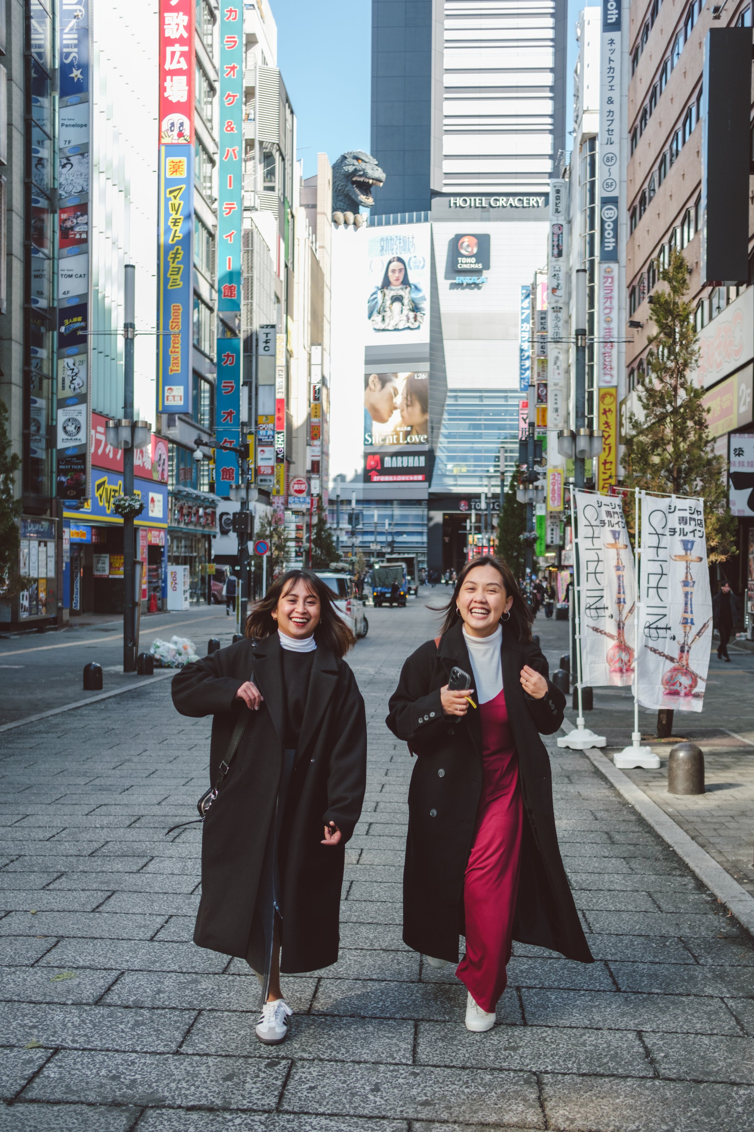 Two women walking down a city street smiling. The street is lined with colorful signs and tall buildings. In the background, there are large billboards and advertisements.