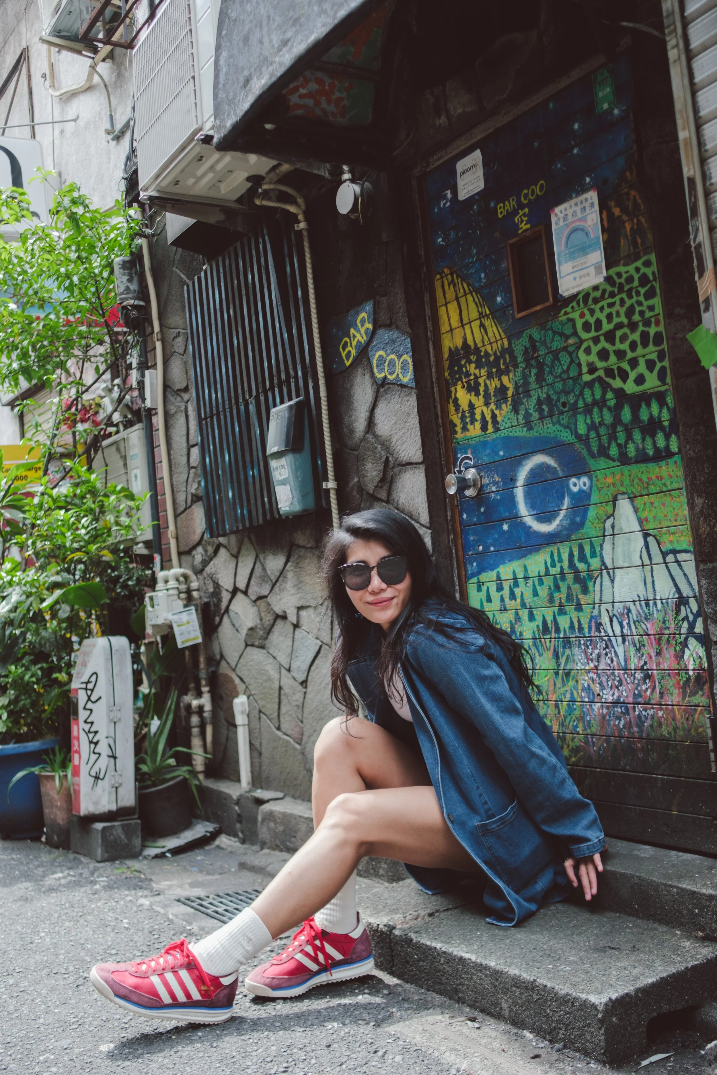Woman sitting on a curb in front of a colorful mural, wearing sunglasses, denim jacket, white socks, and pink sneakers, with urban plants and air conditioning units in the background.