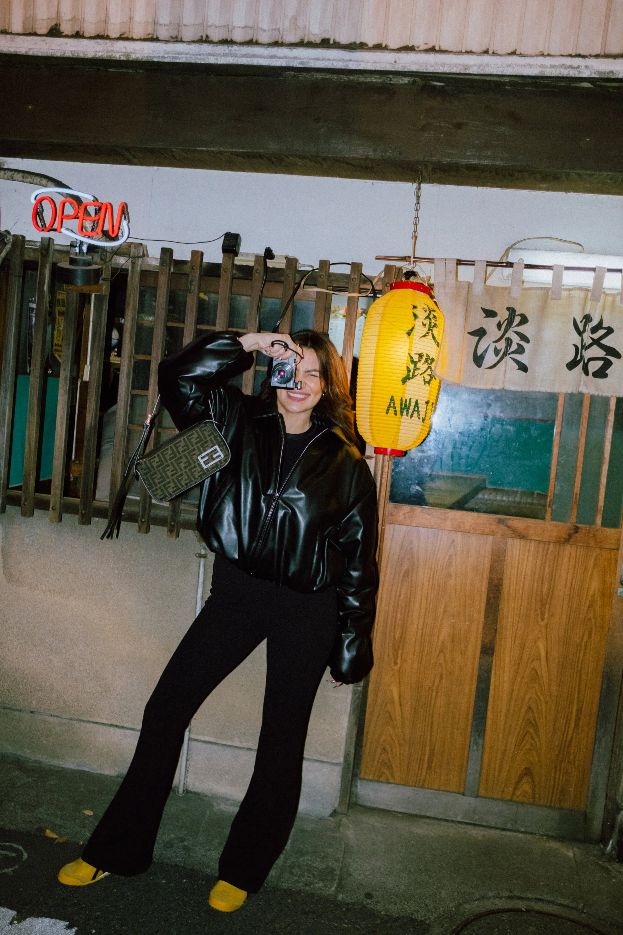 A woman with brown hair smiling and holding a small camera to her eye, standing in front of a wooden structure with a yellow paper lantern hanging nearby that says "AWAJ" and some Japanese characters, at night.
