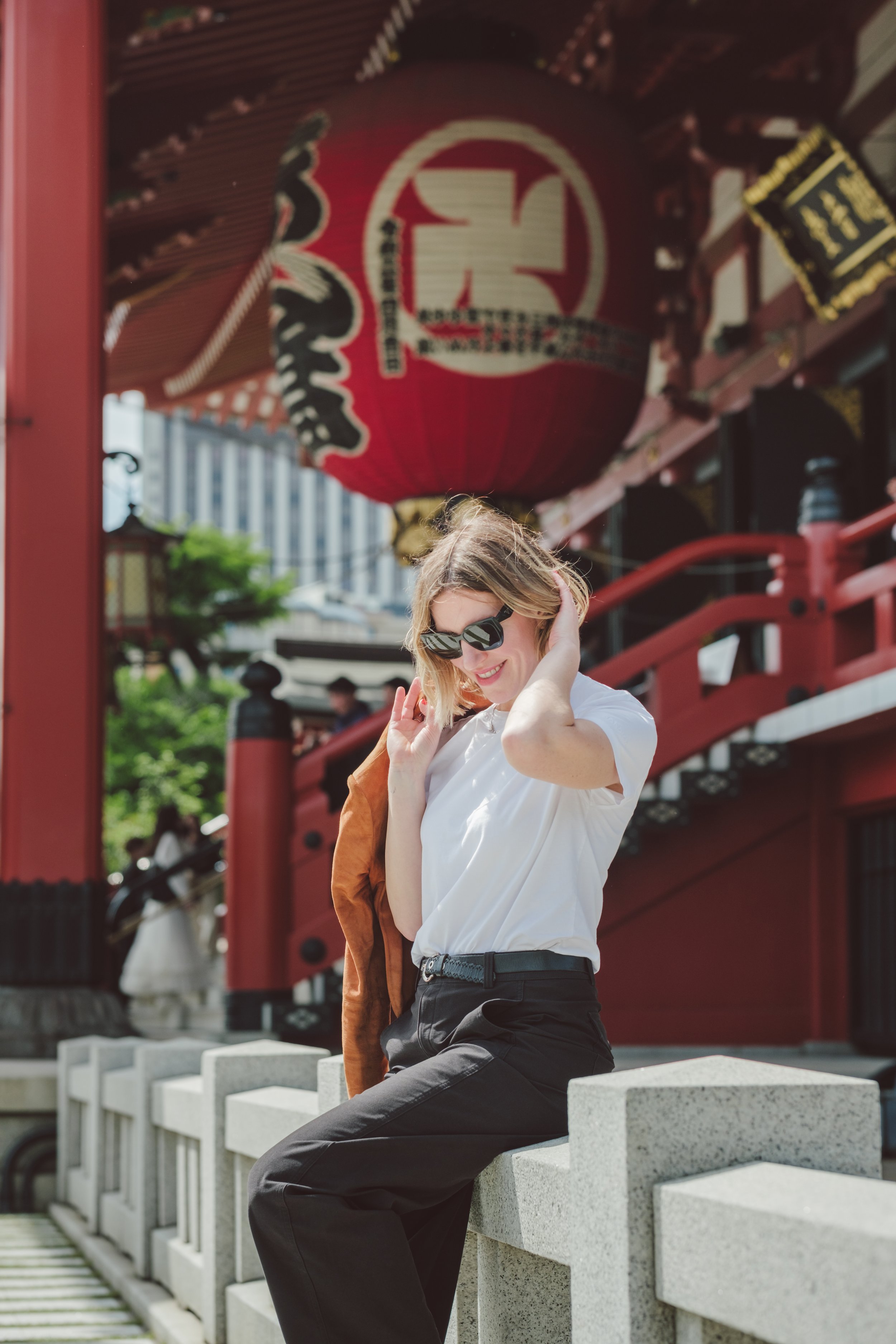 A young woman with blonde hair smiling and wearing sunglasses, a white T-shirt, and black pants, sitting on a concrete barrier in front of a red traditional Japanese temple with a large red lantern hanging overhead.