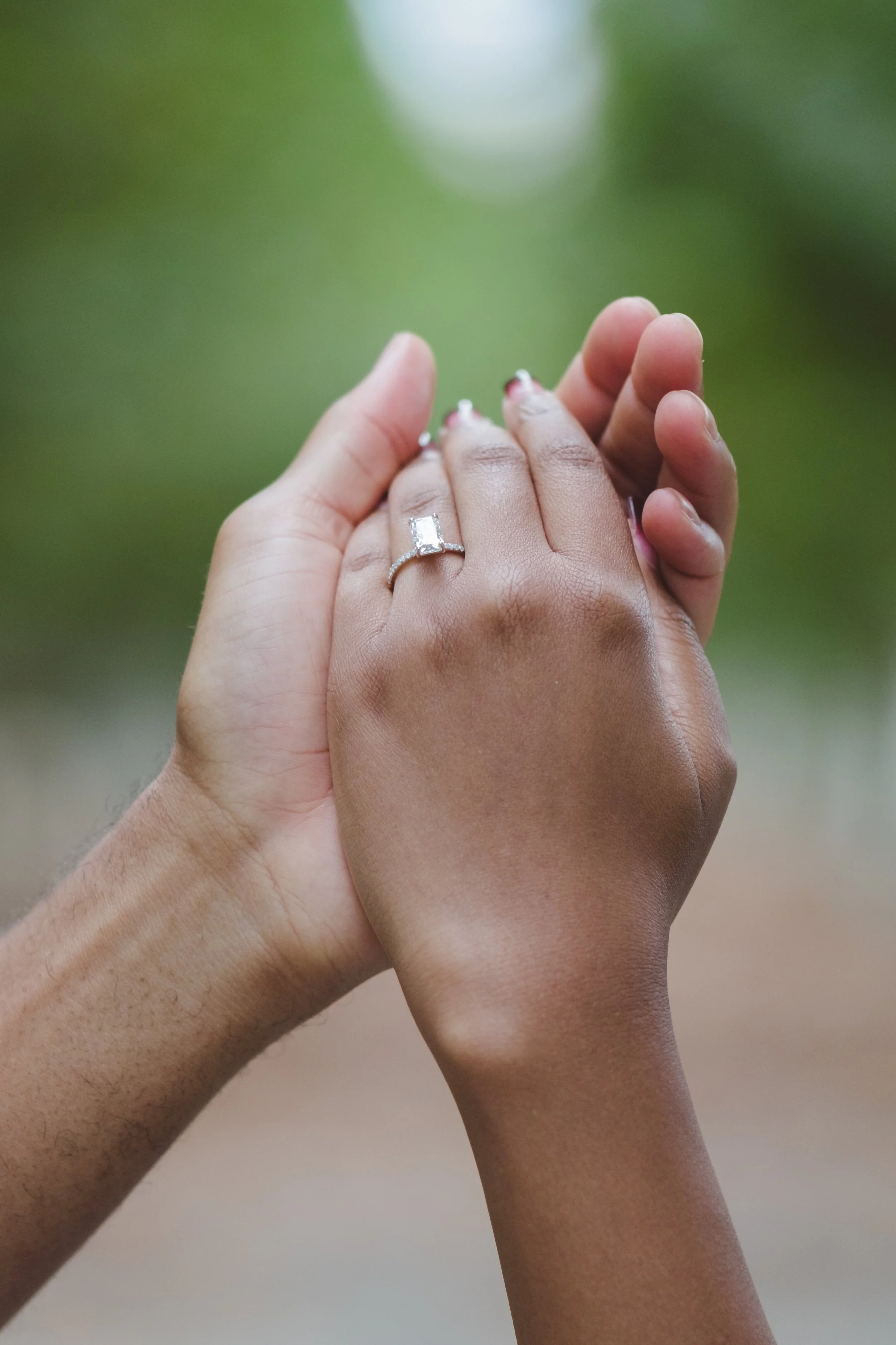 Two hands gently holding each other, with a silver ring featuring a rectangular gemstone on one finger, against a blurred green background.