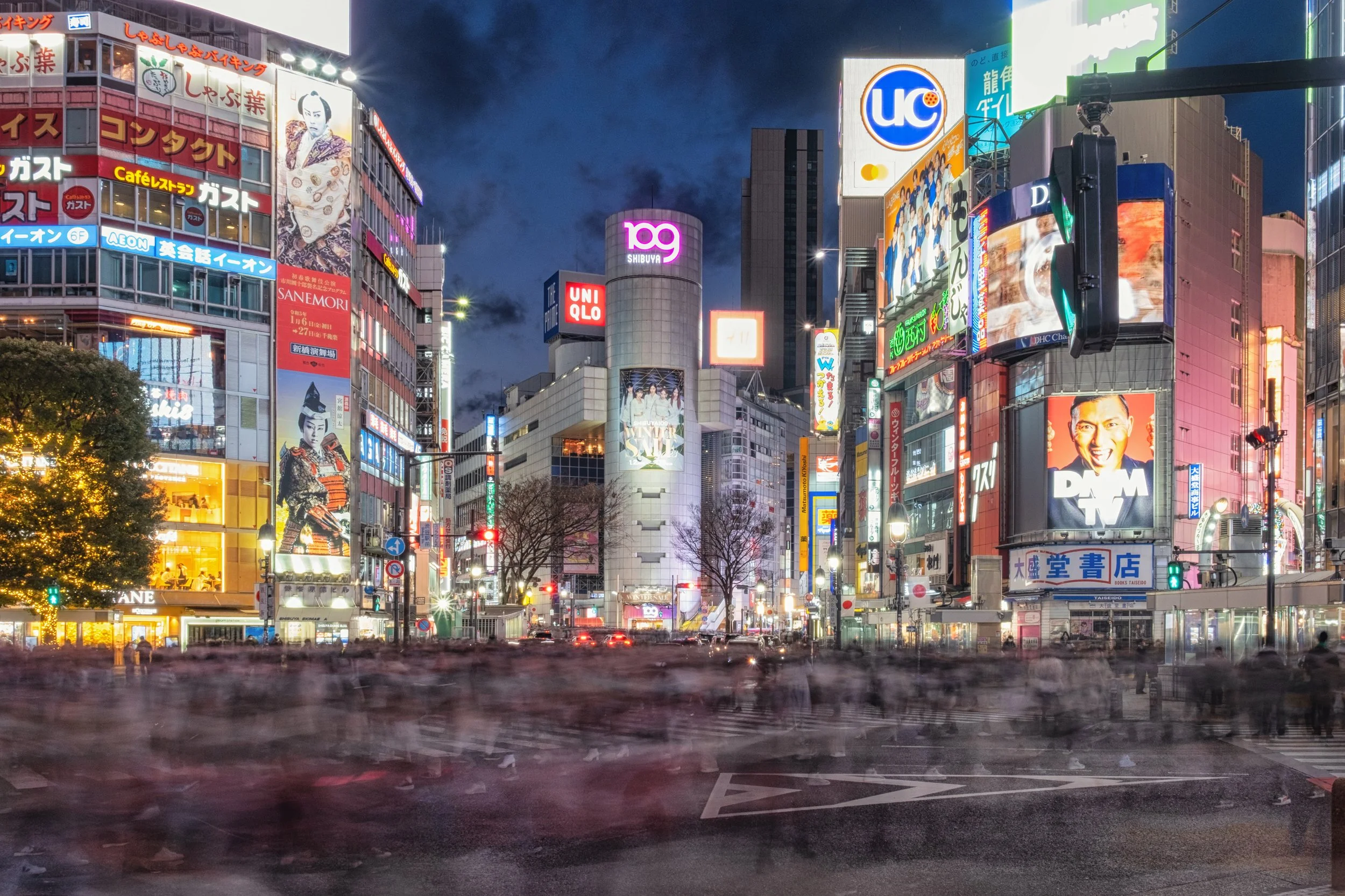 Nighttime cityscape of Shibuya, Tokyo, filled with illuminated billboards and signs on tall buildings, with some blurry cars and pedestrians on the street.