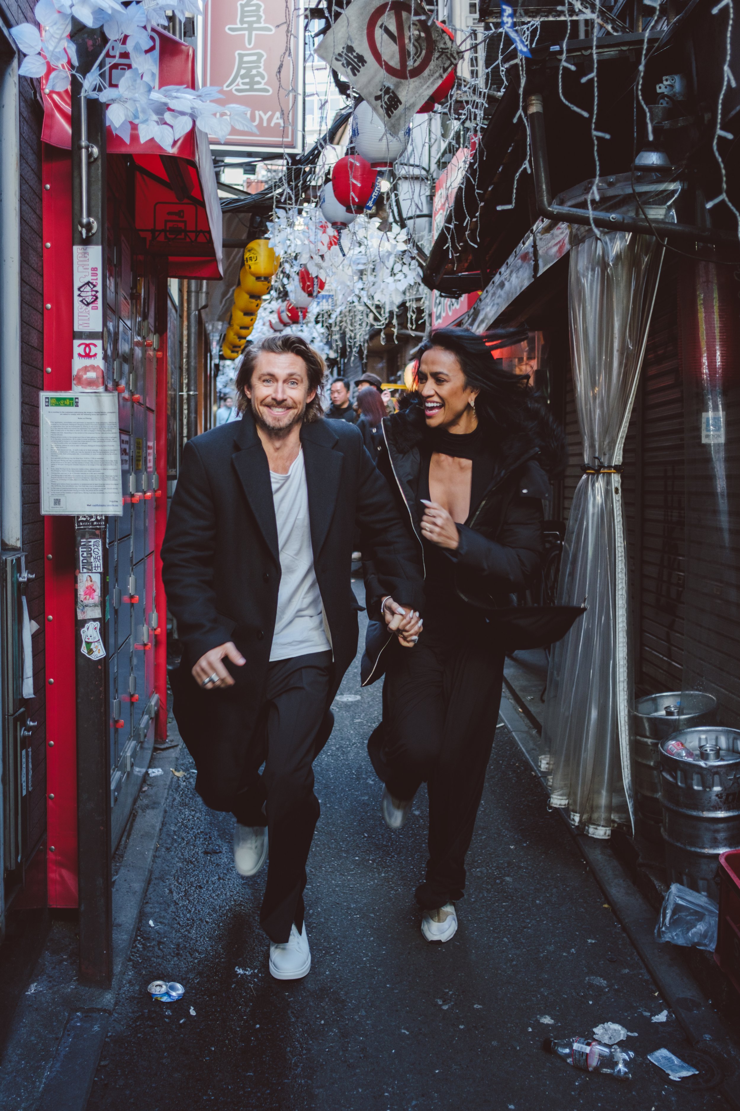 Two people, a man and a woman, walking down a narrow street or alleyway, smiling and holding hands. The street is decorated with hanging lanterns and string lights. There is trash and debris on the ground.