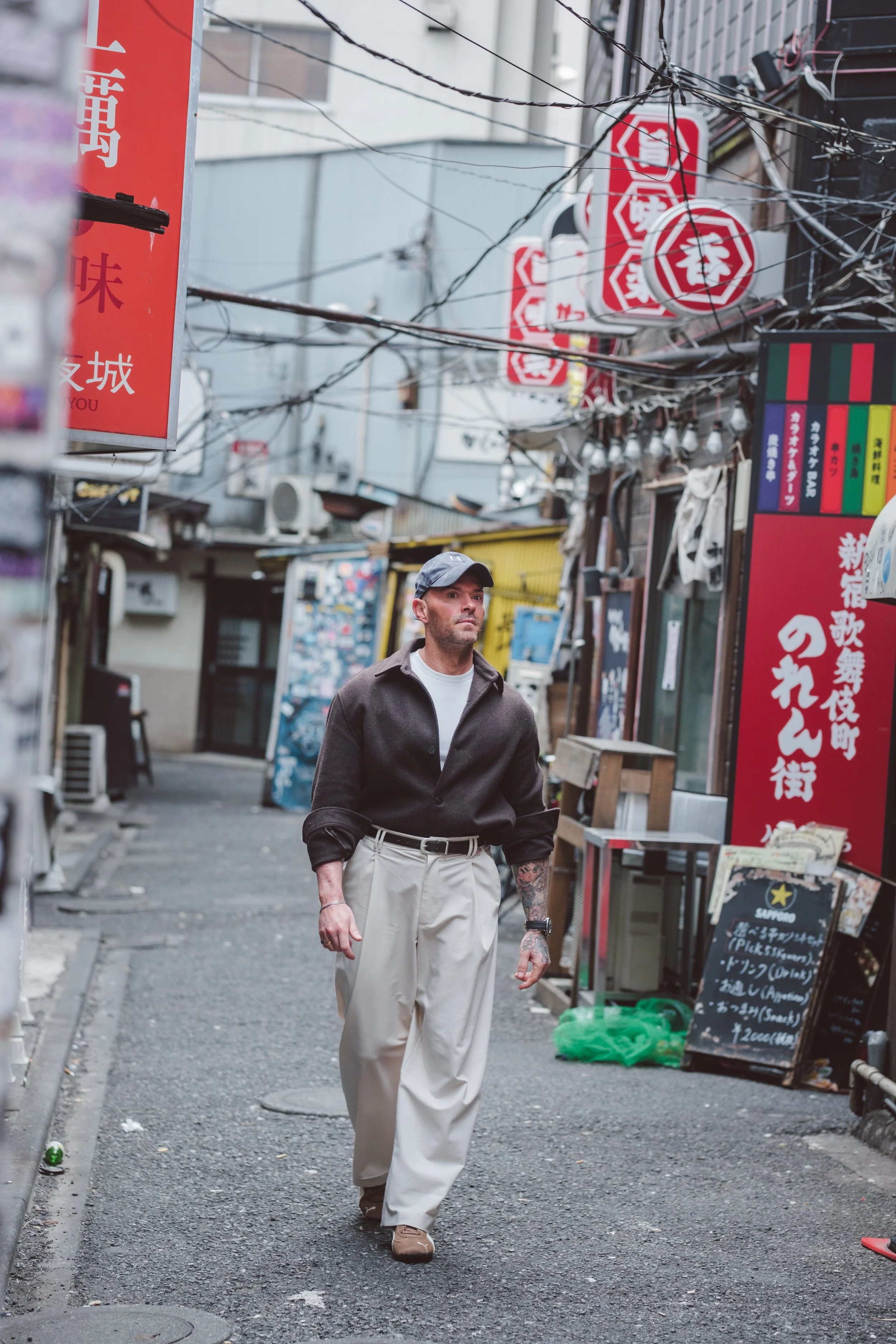 A man in beige pants, a black jacket, and a baseball cap walking down a narrow Asian street alley with signs in Asian languages and shop fronts.