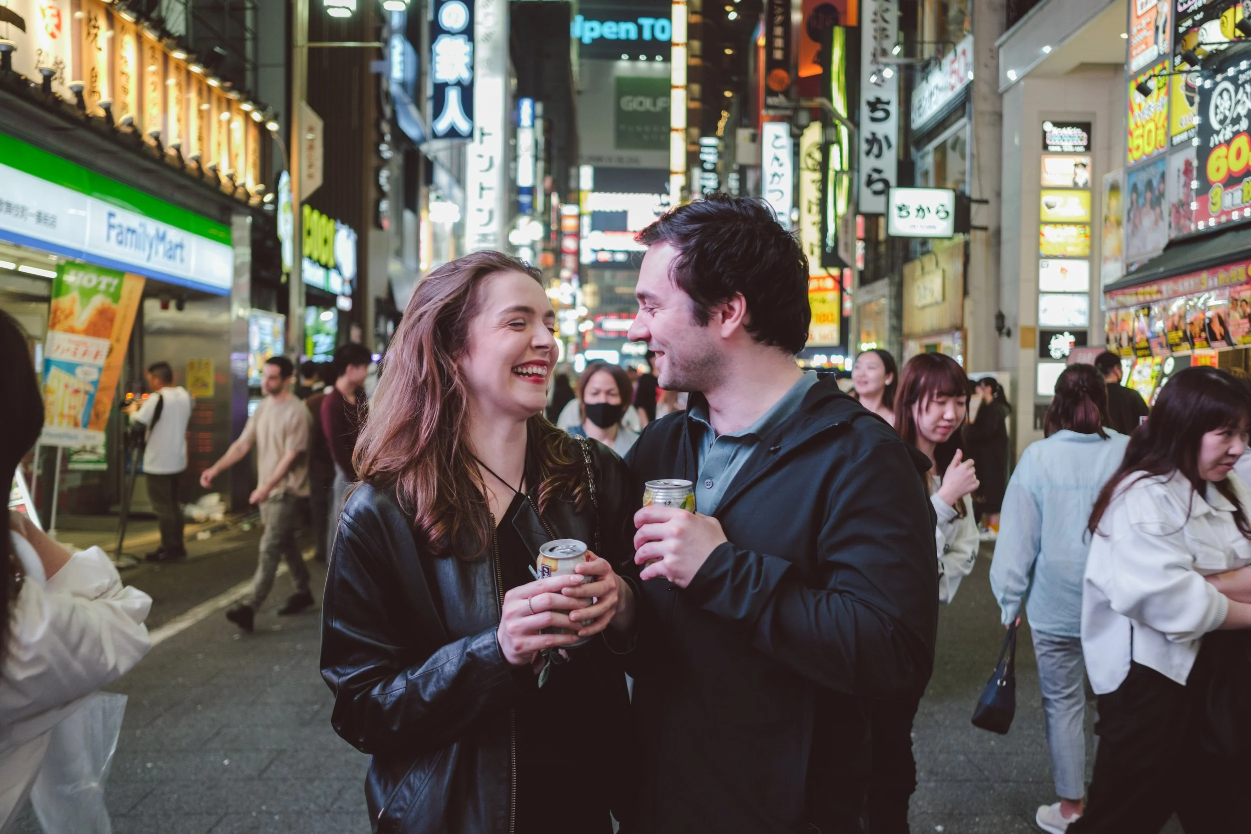 A couple is smiling and looking at each other while holding drinks on a busy city street at night, with bright neon signs and many pedestrians in the background.