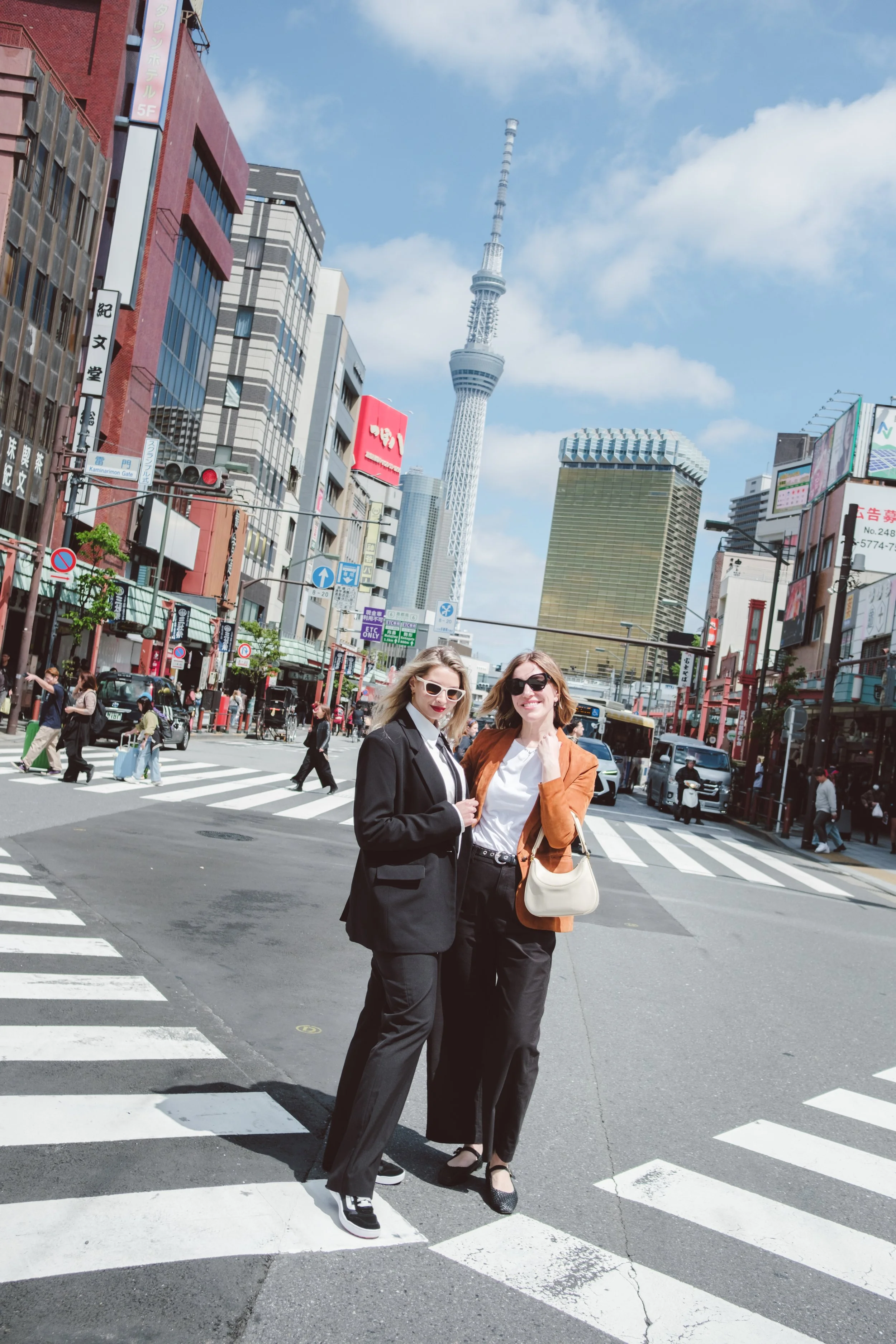 Two women wearing black pants and jackets, smiling and standing close together at a crosswalk in a busy city. The background includes tall buildings, a skyscraper resembling Tokyo Skytree, and a clear blue sky with some clouds.