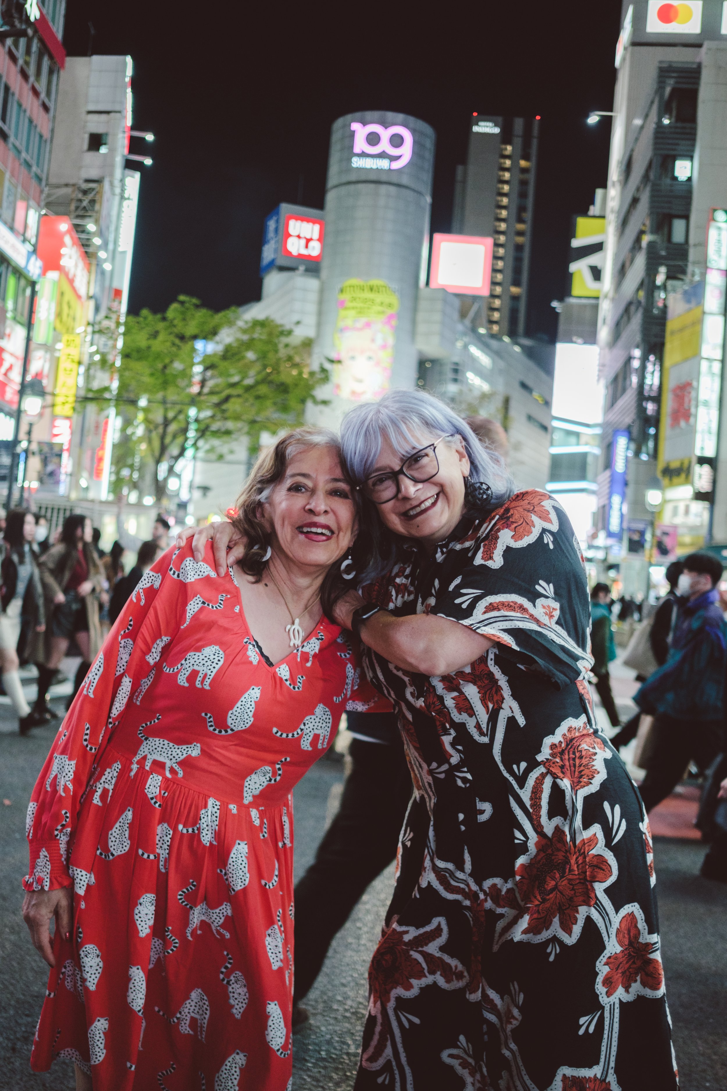 Two women smiling and hugging each other on a busy city street at night, illuminated by bright lights and digital billboards.