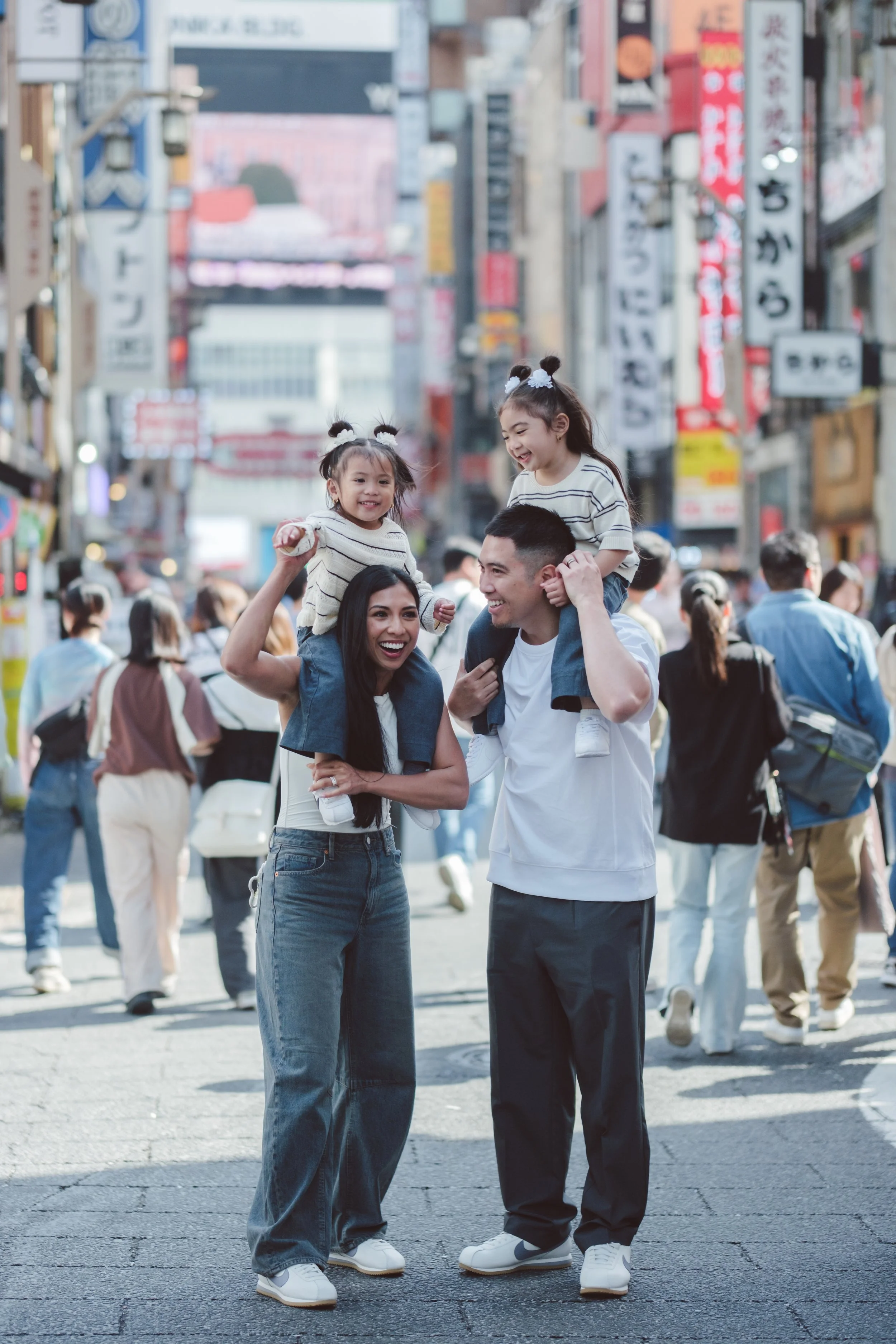 A family of three enjoying a busy urban street, with buildings and signage in the background. The child is sitting on the father's shoulders, and the mother is smiling beside them, all wearing casual clothes and sneakers.