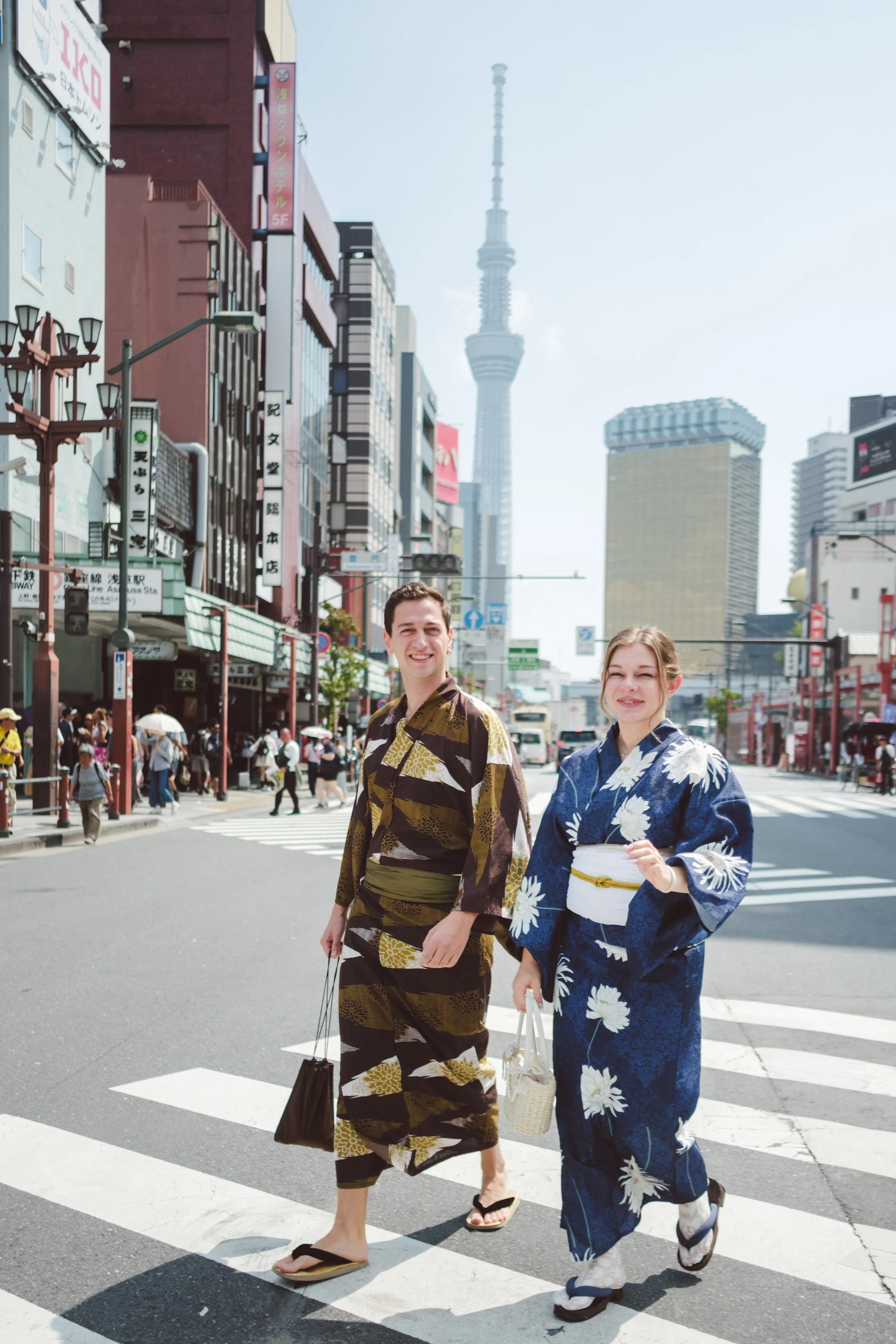 Two people wearing traditional Japanese yukatas walking across a street in a city with modern buildings and skyscrapers, including a tall tower in the background.