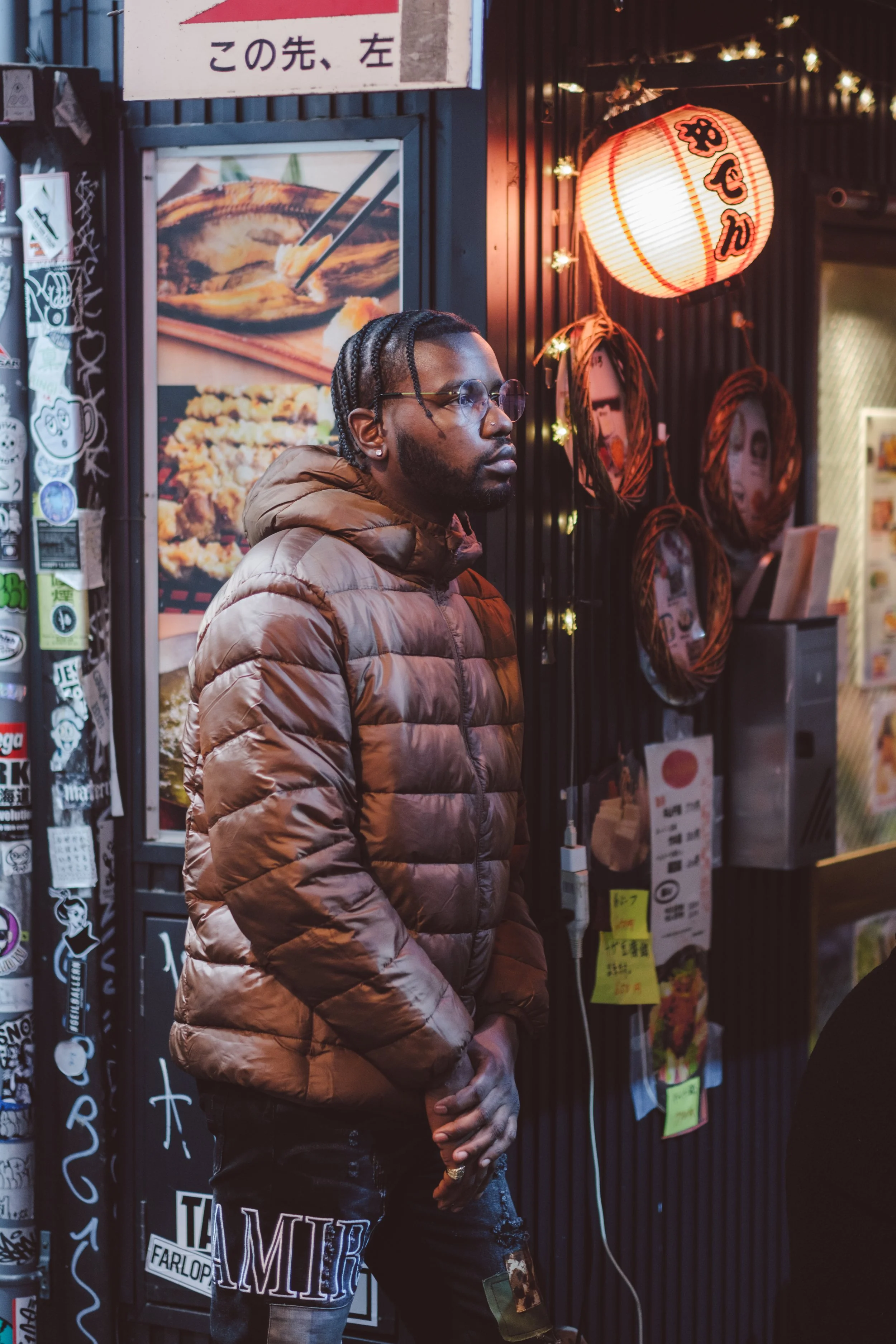 A man with braided hair, glasses, earrings, and a nose piercing, wearing a brown puffer jacket and ripped jeans, stands in front of a Japanese restaurant with paper lanterns and menu displays.