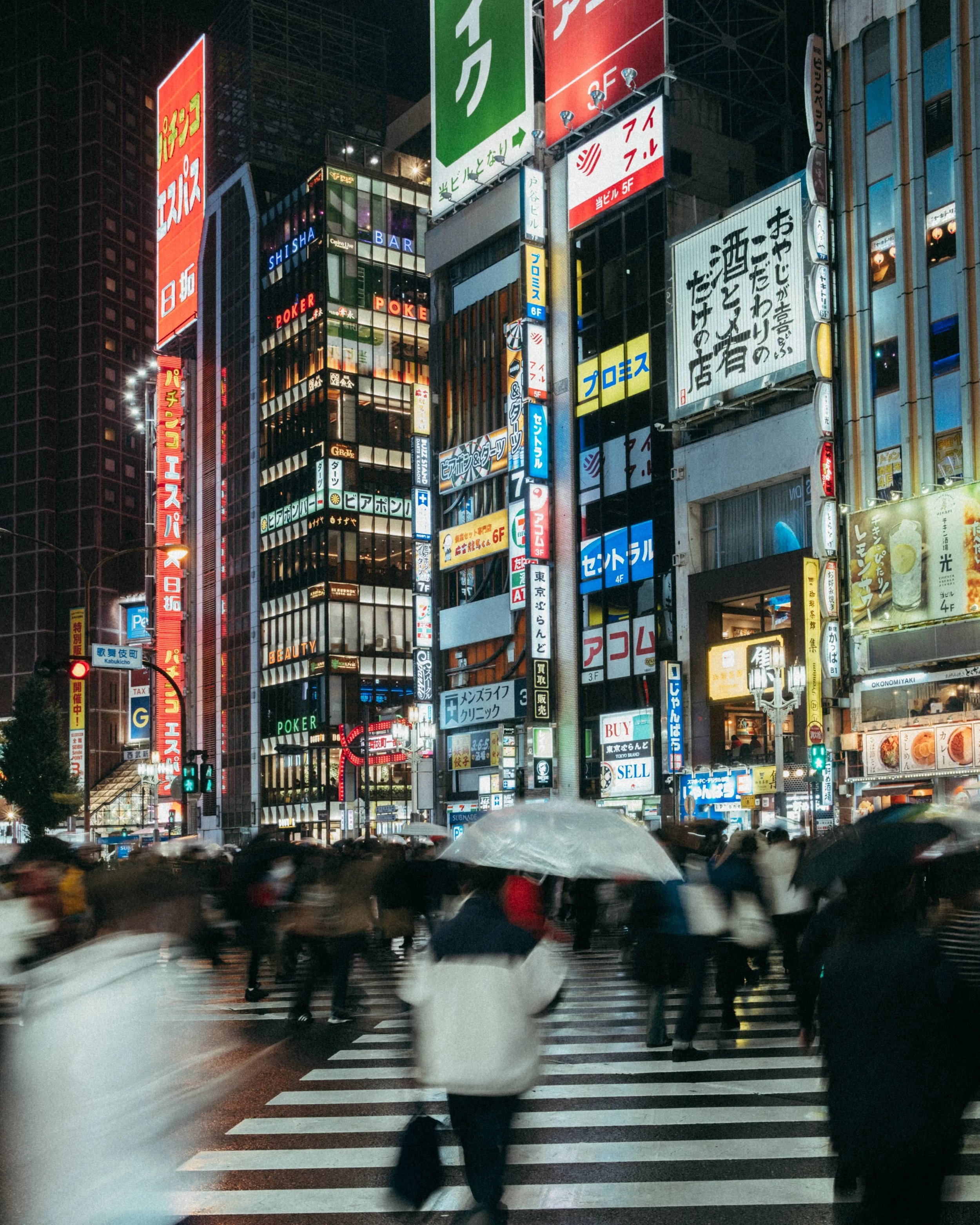 A city street at night with neon signs and a crowd crossing the street under umbrellas in rainy weather.