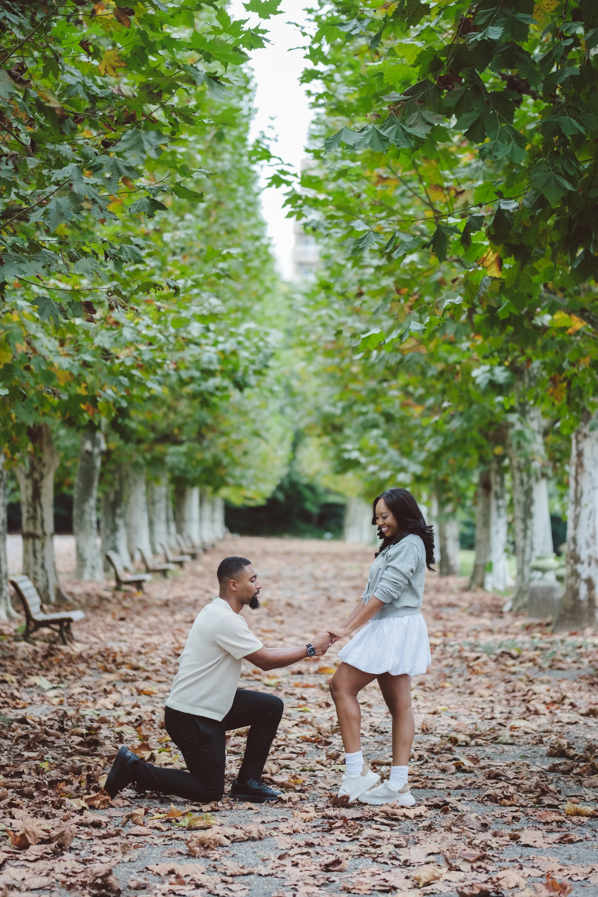 A man proposing marriage to a woman in a park with fallen autumn leaves and trees with green leaves.