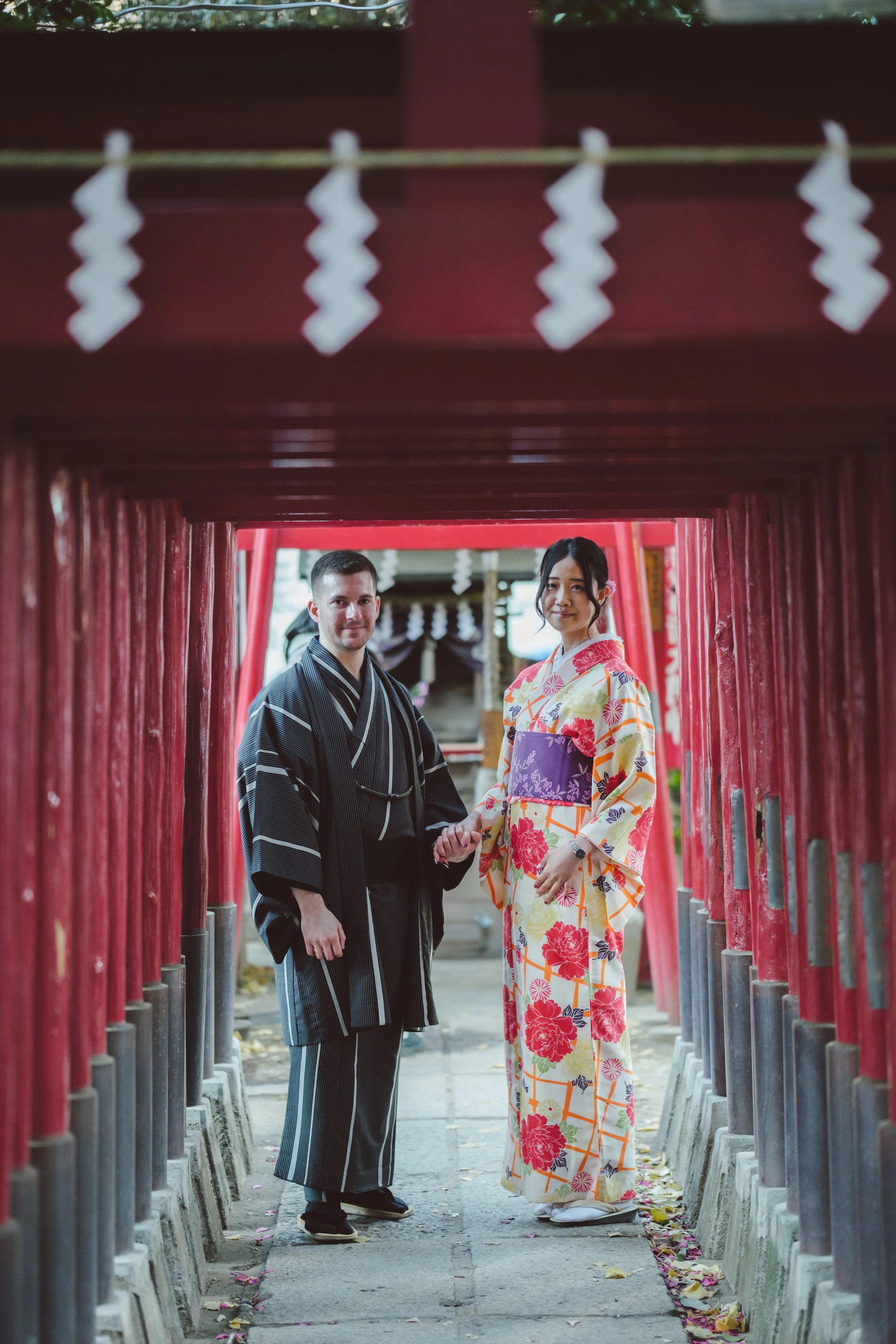 A couple dressed in traditional Japanese kimonos holding hands under a red torii gate at a shrine.