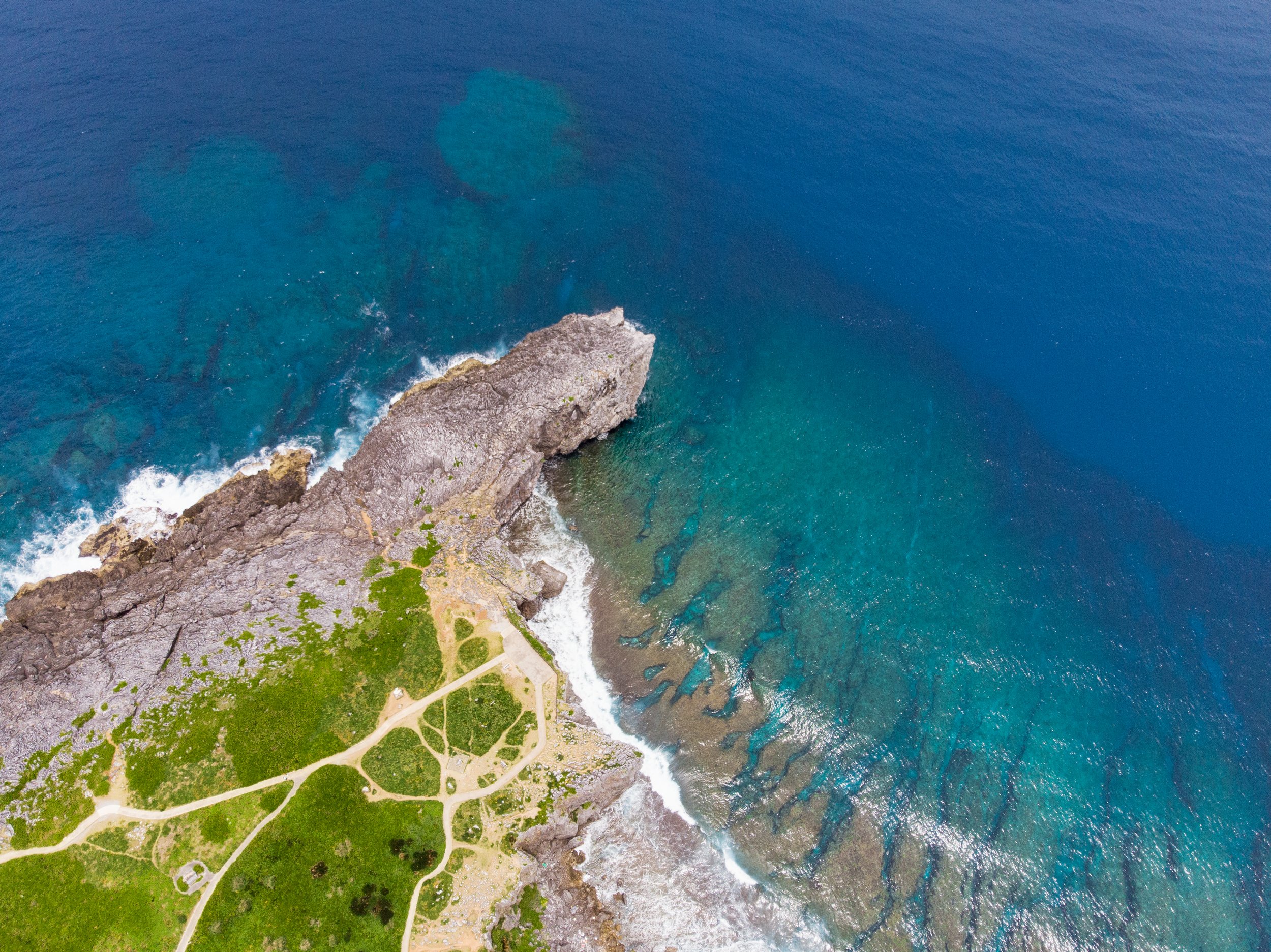 Aerial view of a coastline with rocky cliffs, green vegetation, and a pathway leading to the water, with blue ocean waves crashing against the rocks.