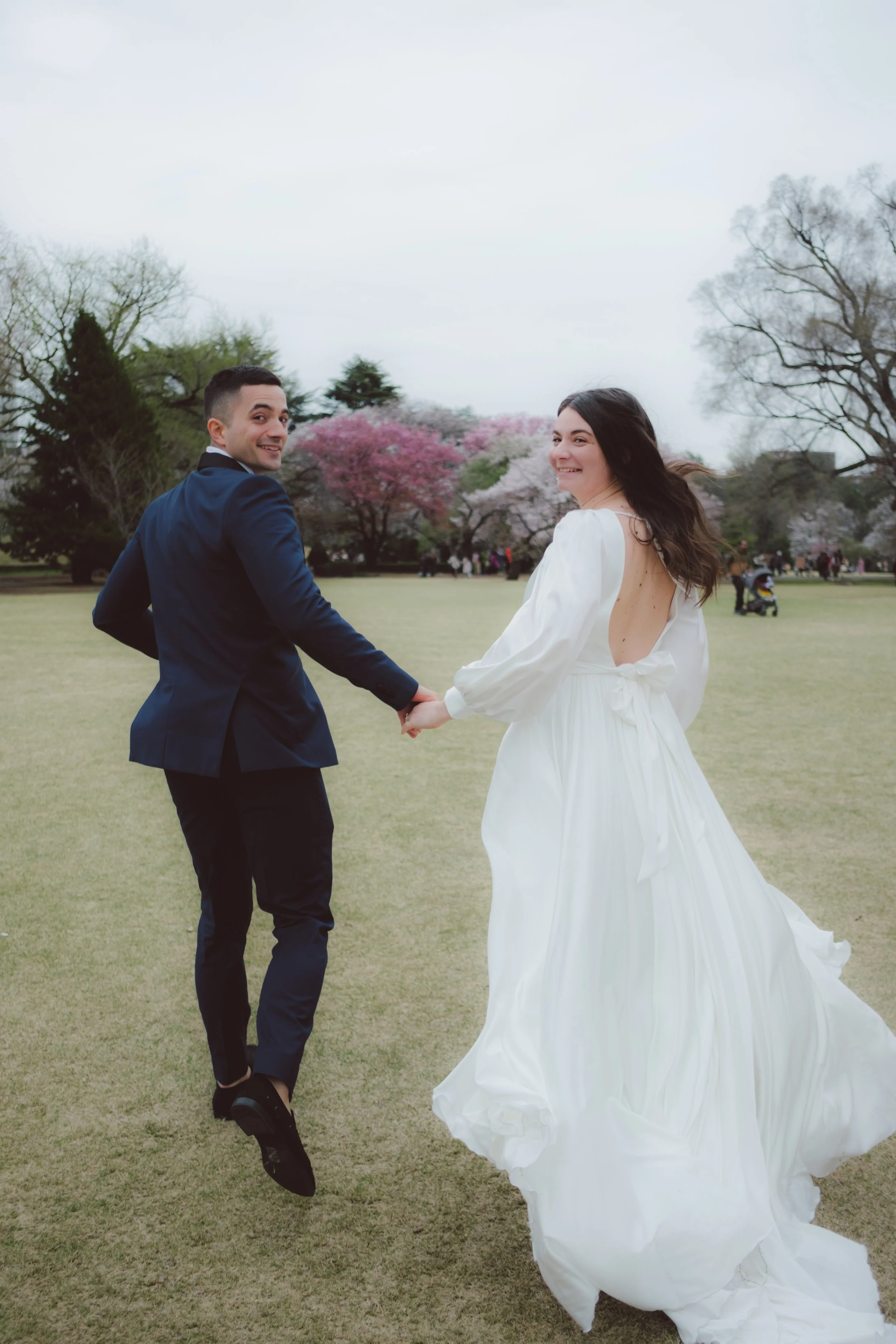 A bride and groom holding hands and spinning outdoors in a park with blooming cherry blossom trees, smiling and looking back at the camera.