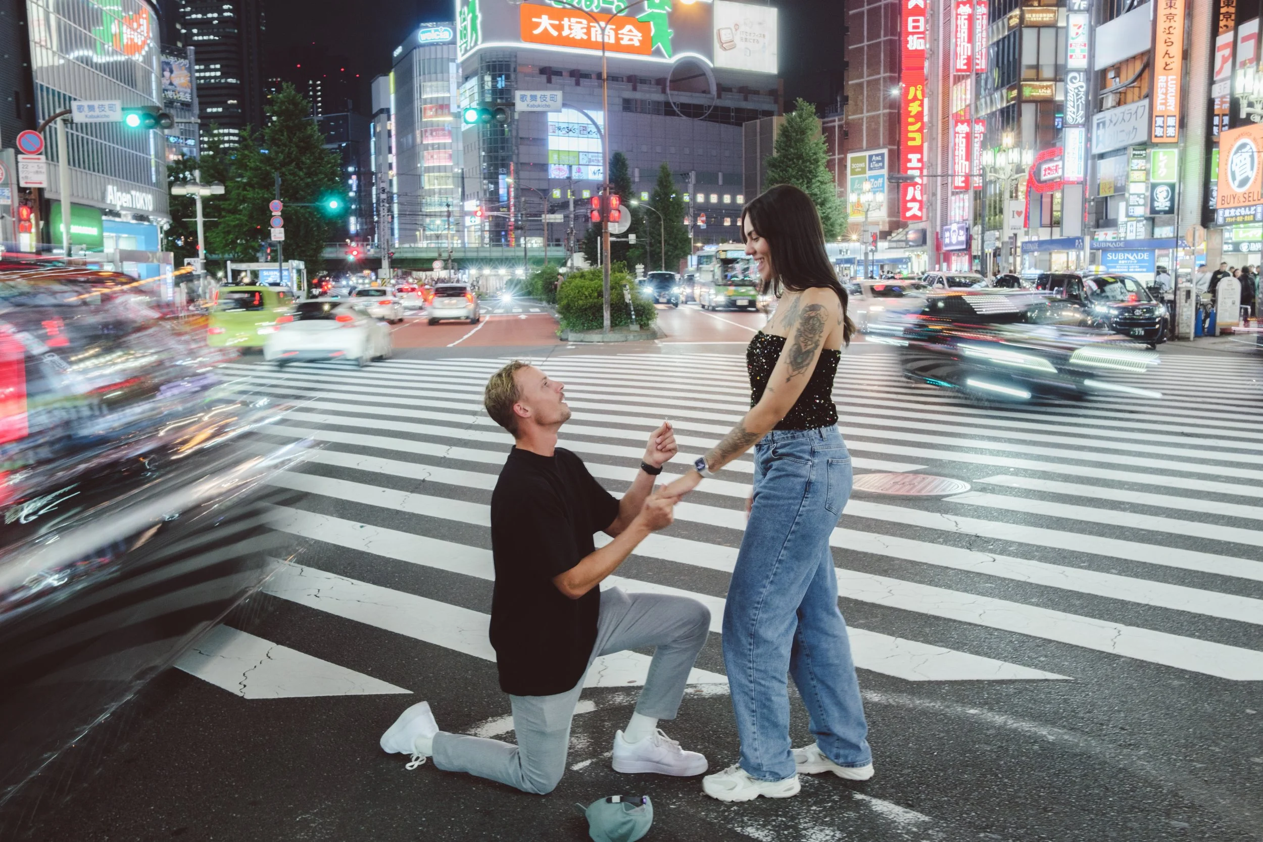 A man kneeling on the crosswalk holding a woman's hand, who is standing and smiling, in a busy city at night with bright neon signs and cars passing by.