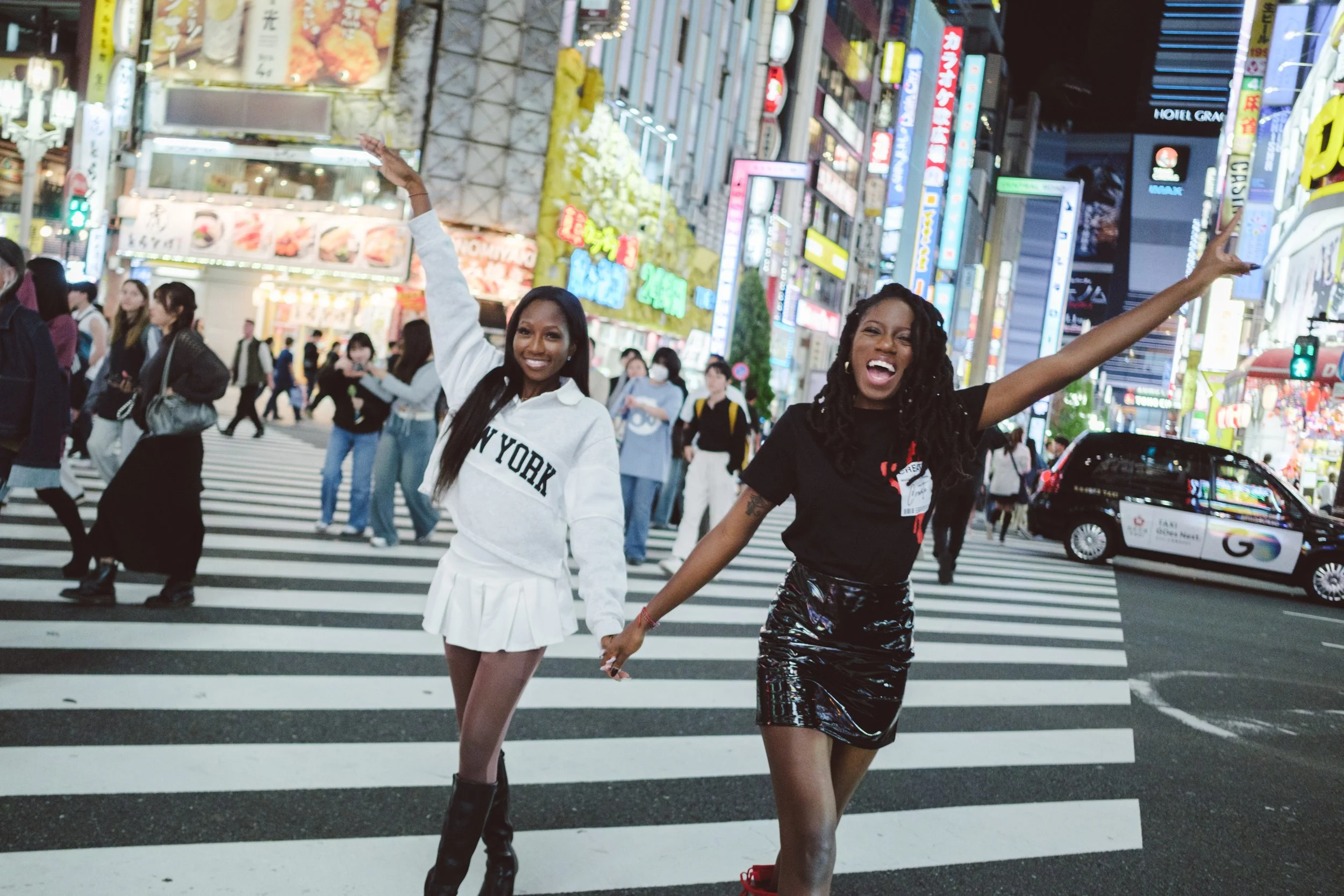 Two women holding hands, dancing or celebrating in the crosswalk of a busy city street at night, surrounded by illuminated signs and people.