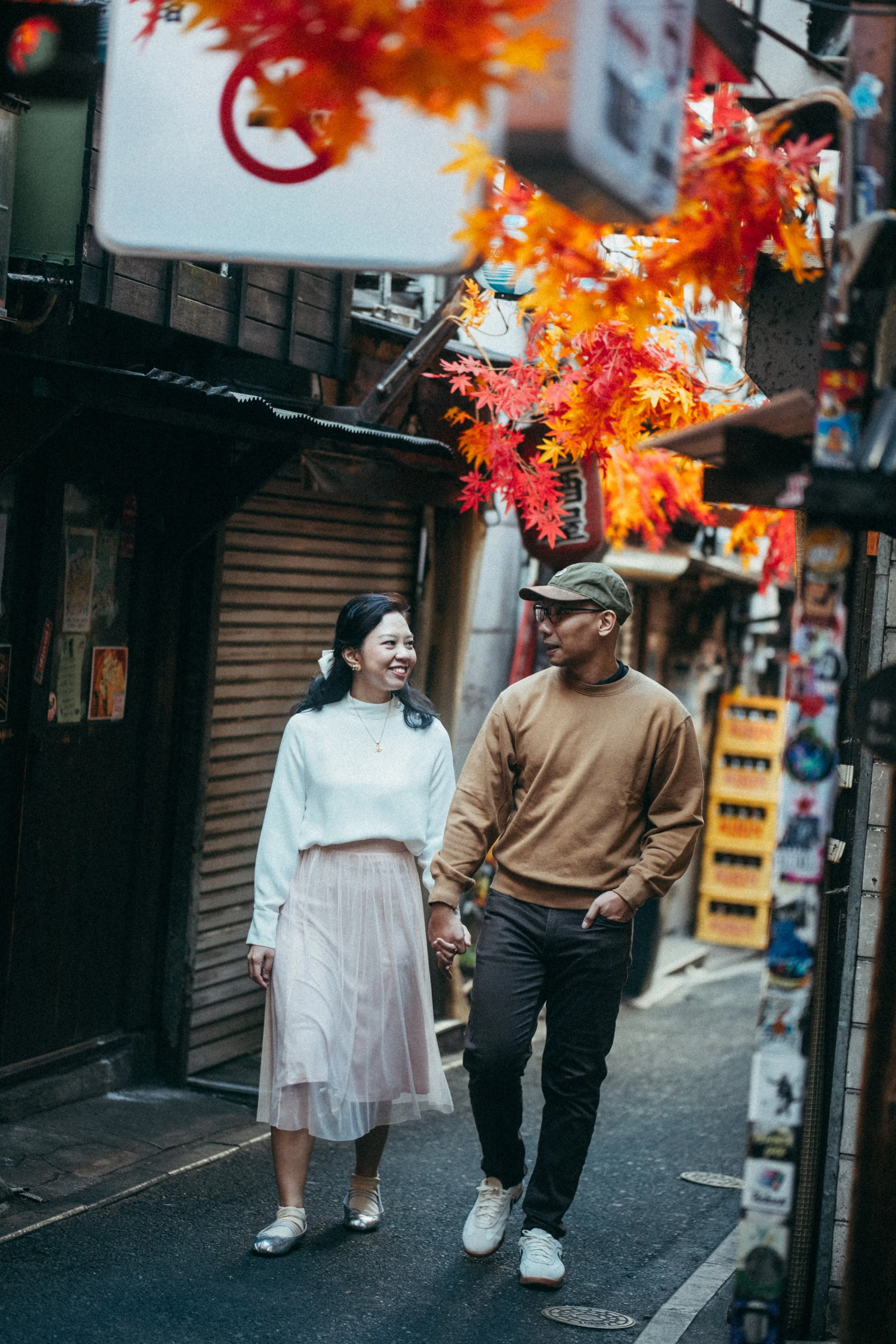 A young couple walking hand in hand in an outdoor urban setting decorated with autumn leaves. They are smiling and engaged in conversation.