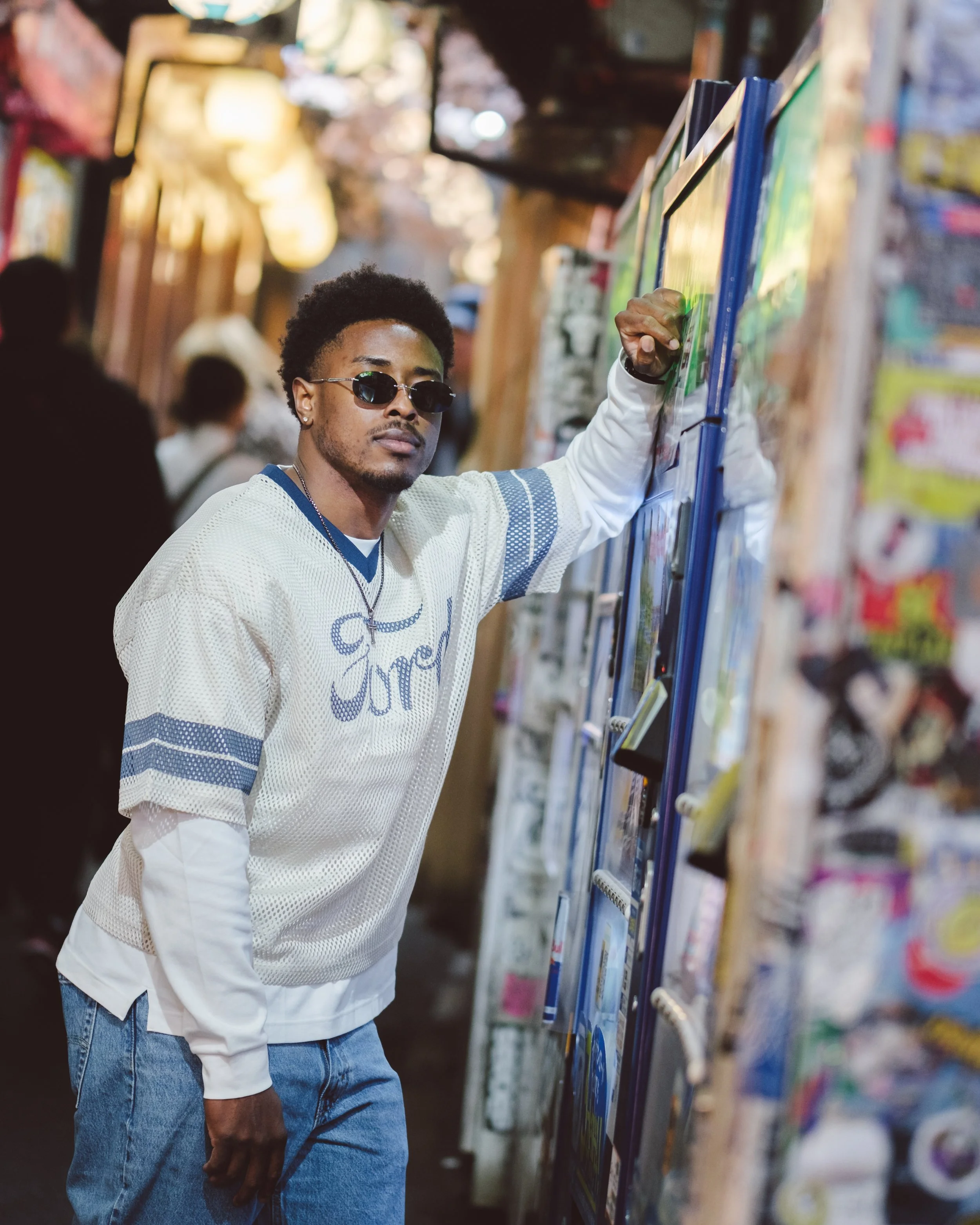 A young man with curly hair wearing sunglasses, a white sweatshirt with text, and jeans, shopping at a store. He is looking at items on a display shelf in a store aisle with bright, warm lighting.