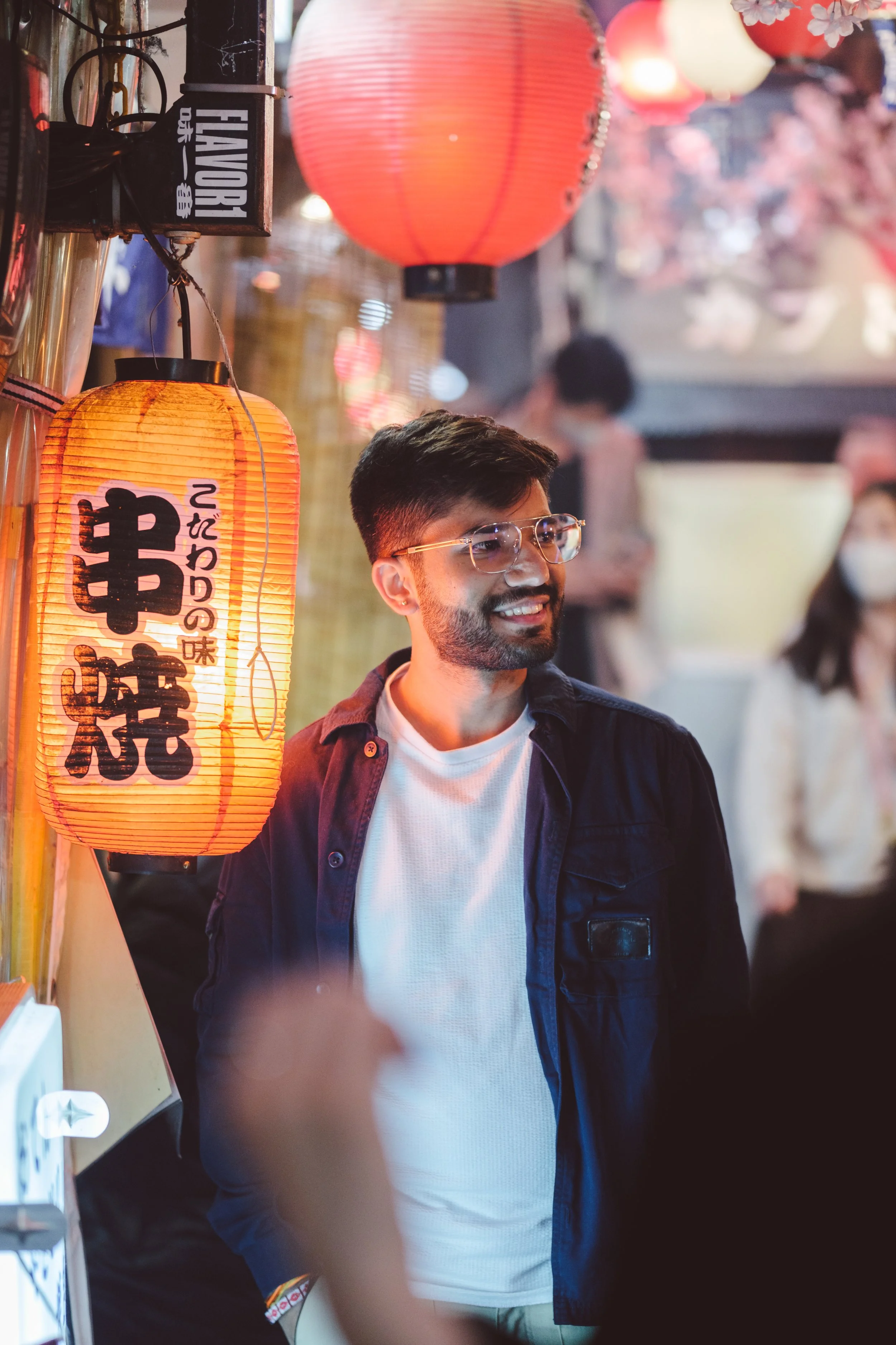 A man with glasses and a beard smiling, standing in front of Japanese lanterns. There are other people in the background, some wearing masks, in a lively outdoor setting.