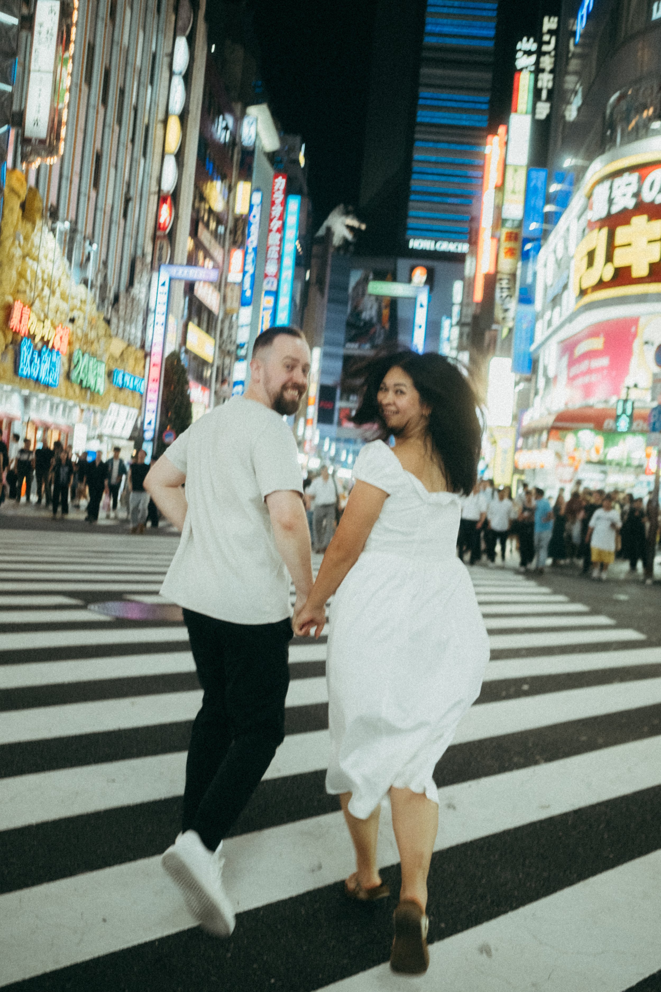 A man and a woman holding hands and walking on a crosswalk at night in a busy city, illuminated by colorful neon signs and billboards.