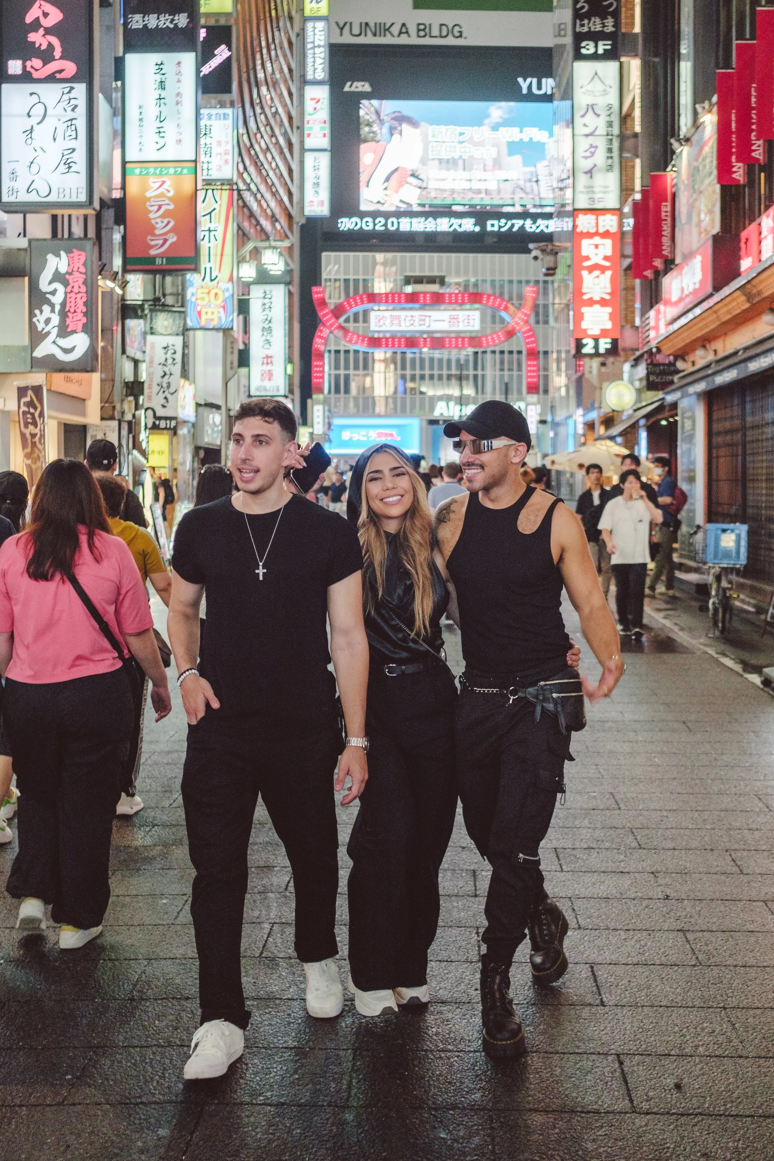 Three young people walking together in a busy city street at night, with neon signs and billboards in the background. Two men and one woman are smiling and dressed in casual dark clothing.