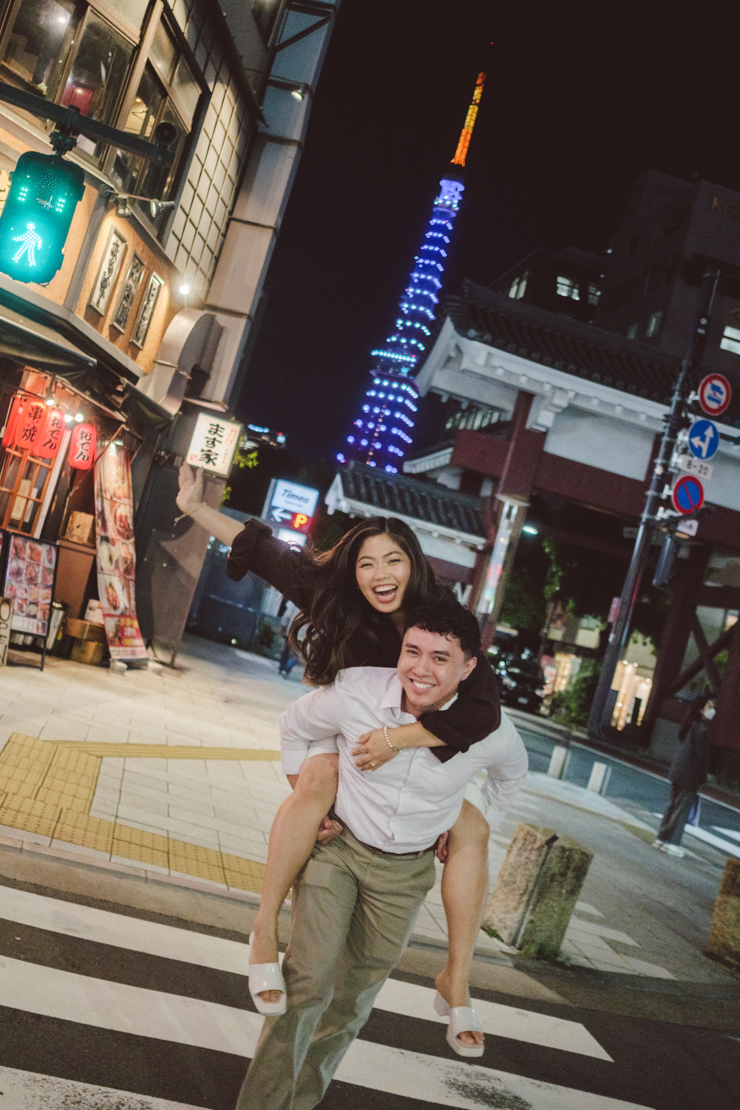 A night scene on a city street shows a man carrying a woman on his back, both smiling and laughing. In the background, the Tokyo Tower is lit up with blue and orange lights. Surrounding buildings include restaurants and shops with colorful signs, inc