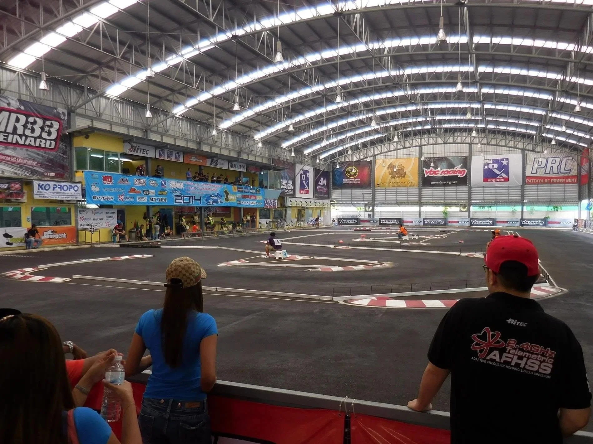 Indoor RC car racing track with a curved layout, surrounded by spectators. The venue has a high arched ceiling with skylights, and there are banners and advertisements on the walls.
