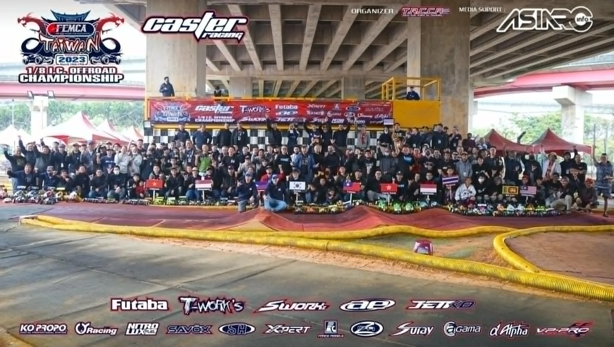 Group photo of participants and officials at the Taiwan 2023 Offroad Championship, taken under a concrete bridge, with banners and logos of sponsors and organizers visible. People are standing and sitting in front of a dirt racing track, with some holding flags and trophies.