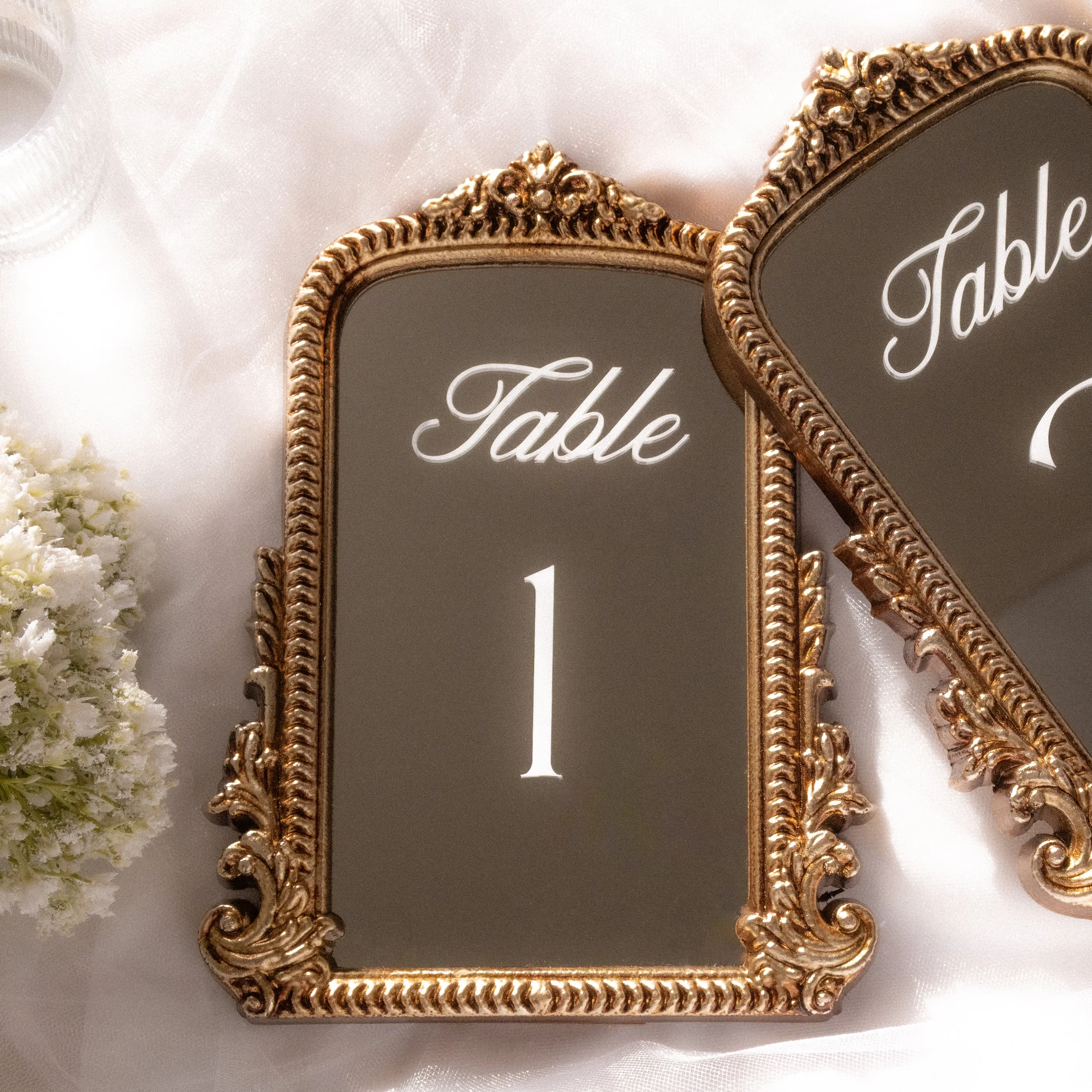 Decorative gold-framed mirror with the words 'Table' and a number 1 on the mirror surface, placed on a white satin cloth with a white floral arrangement nearby.