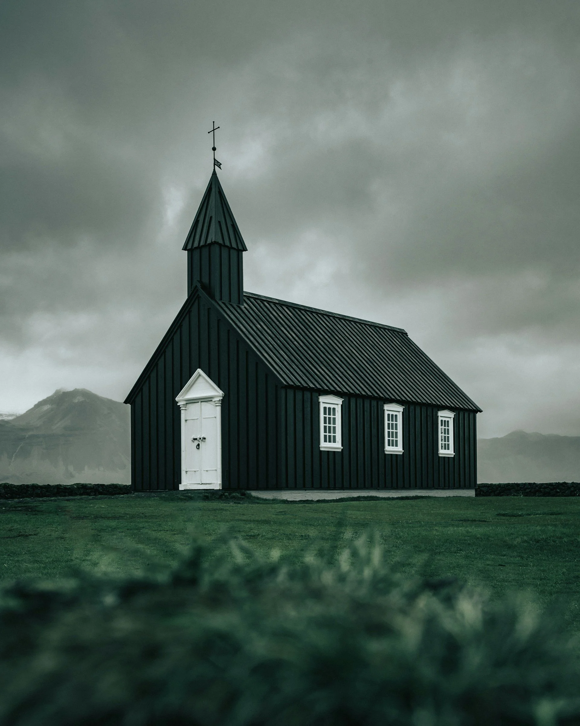 A small black wooden church with white doors and window trim under a cloudy sky, with mountains in the background and green grass in the foreground.