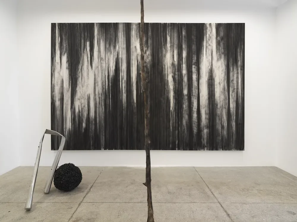 An art gallery with a large black and white abstract painting on a white wall, a curved metal sculpture, a black textured sculpture, and a thin vertical wooden branch in the foreground.