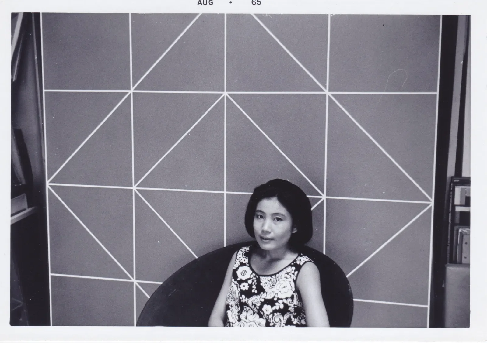 A woman with short dark hair wearing a sleeveless floral dress, sitting on a curved chair in front of a geometric patterned wall with intersecting lines, in a black and white photo.