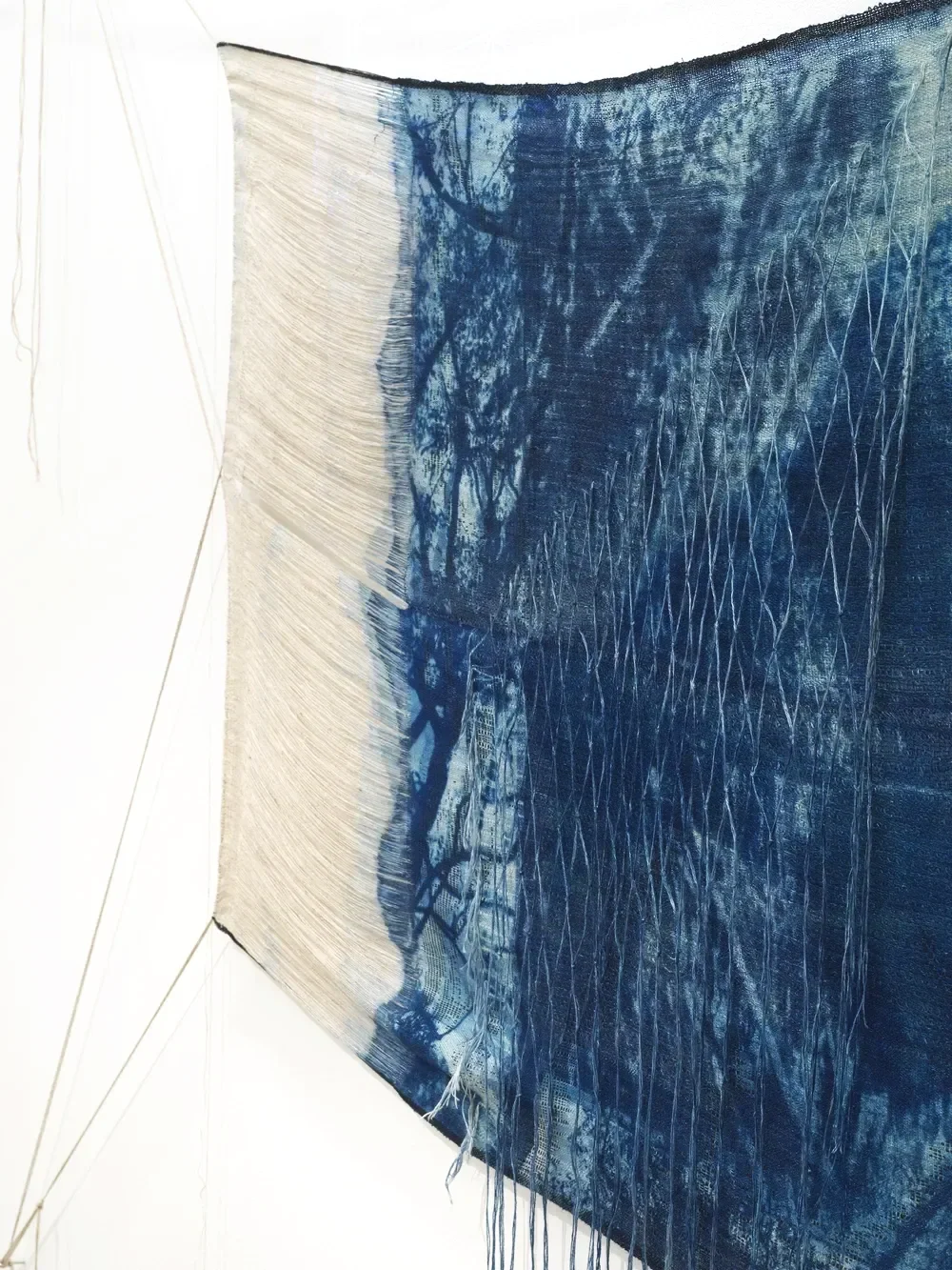 A textile art piece hanging on a white wall, consisting of a large fabric with a printed landscape scene in shades of blue and white, with frayed edges and embroidered lines.
