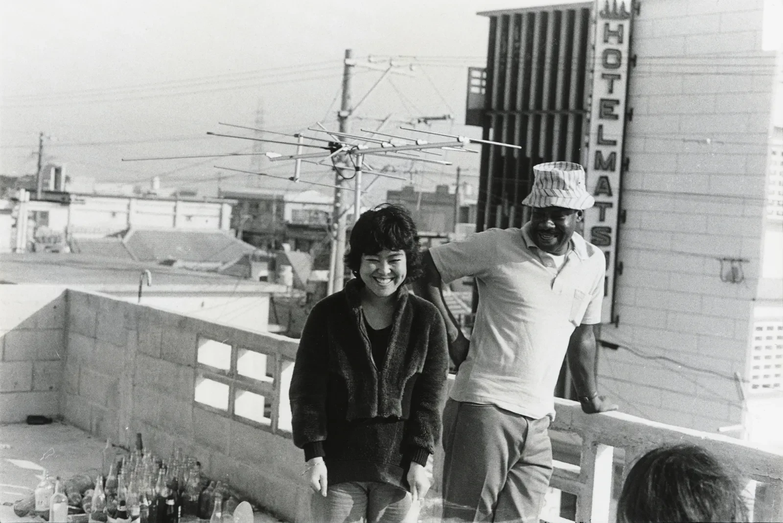 A black and white photo of a smiling woman and man standing outdoors on a rooftop, with buildings and power lines in the background. The man has his arm around the woman and is wearing a bucket hat and a light-colored shirt.