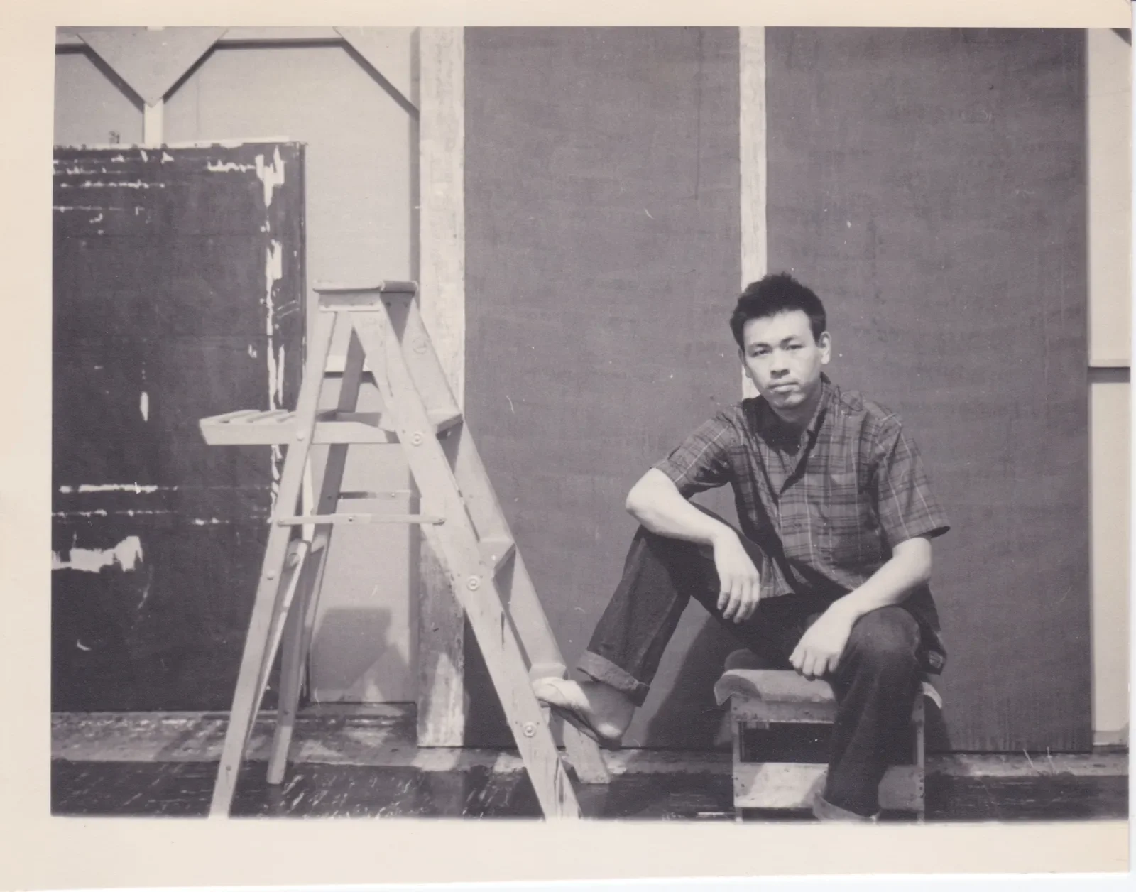 Black and white photo of a young man in a plaid shirt sitting on a stool next to a ladder in a room with plain walls, possibly in a studio or workspace.