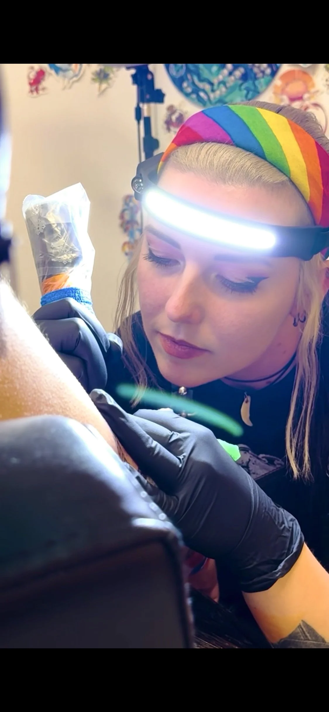Close-up of a woman with rainbow headband, and wearing black gloves, giving a tattoo to a client. A tattoo needle, tattoo ink, and tattoo equipment visible.