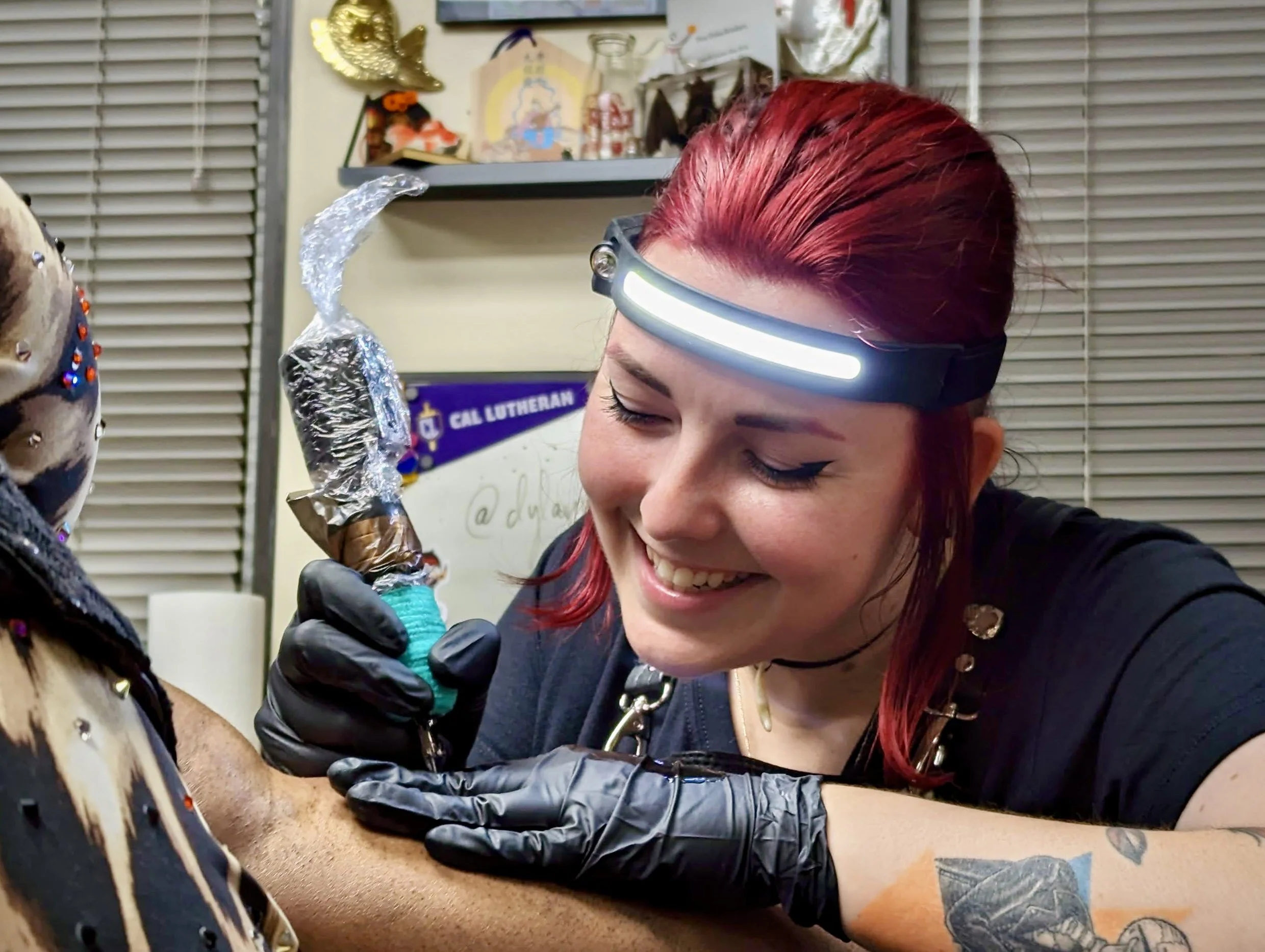 Woman with red hair and a headlamp tattooing a person's arm during a tattoo session.
