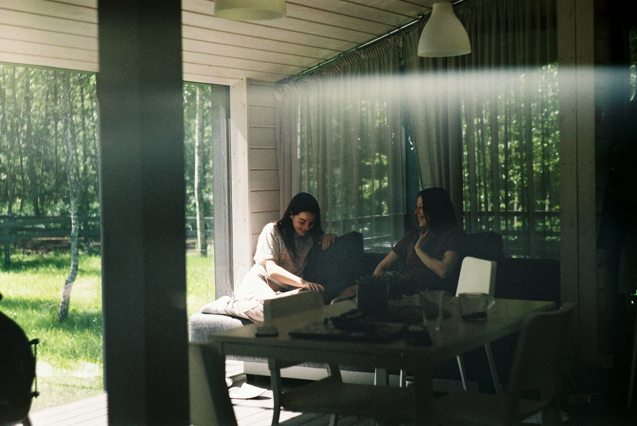 Two women sitting together indoors, smiling and talking, with large windows showing a green outdoor area with trees and grass.