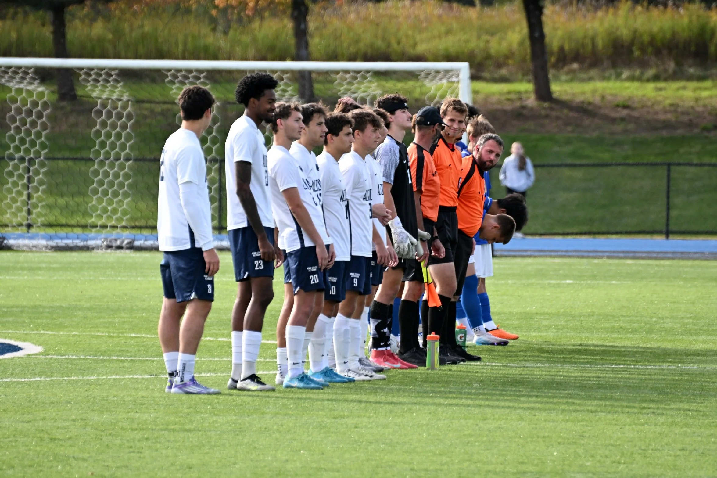 A group of soccer players and referees lined up on the field before a game, with trees in the background.