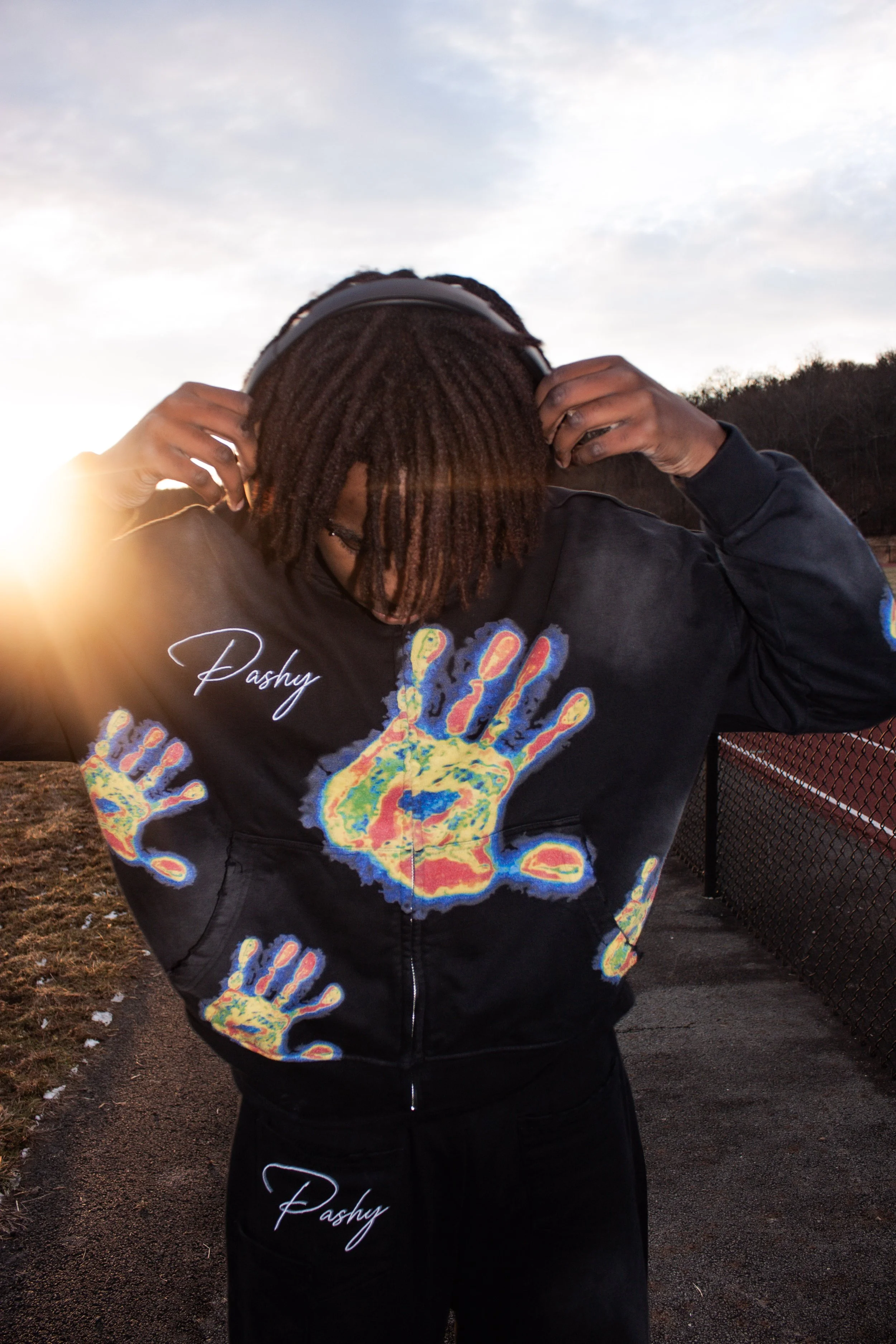 Person with dreadlocks wearing a black hoodie with colorful handprints and the word 'Pashy' on it, adjusting headphones outdoors during sunset with a chain-link fence and trees in the background.