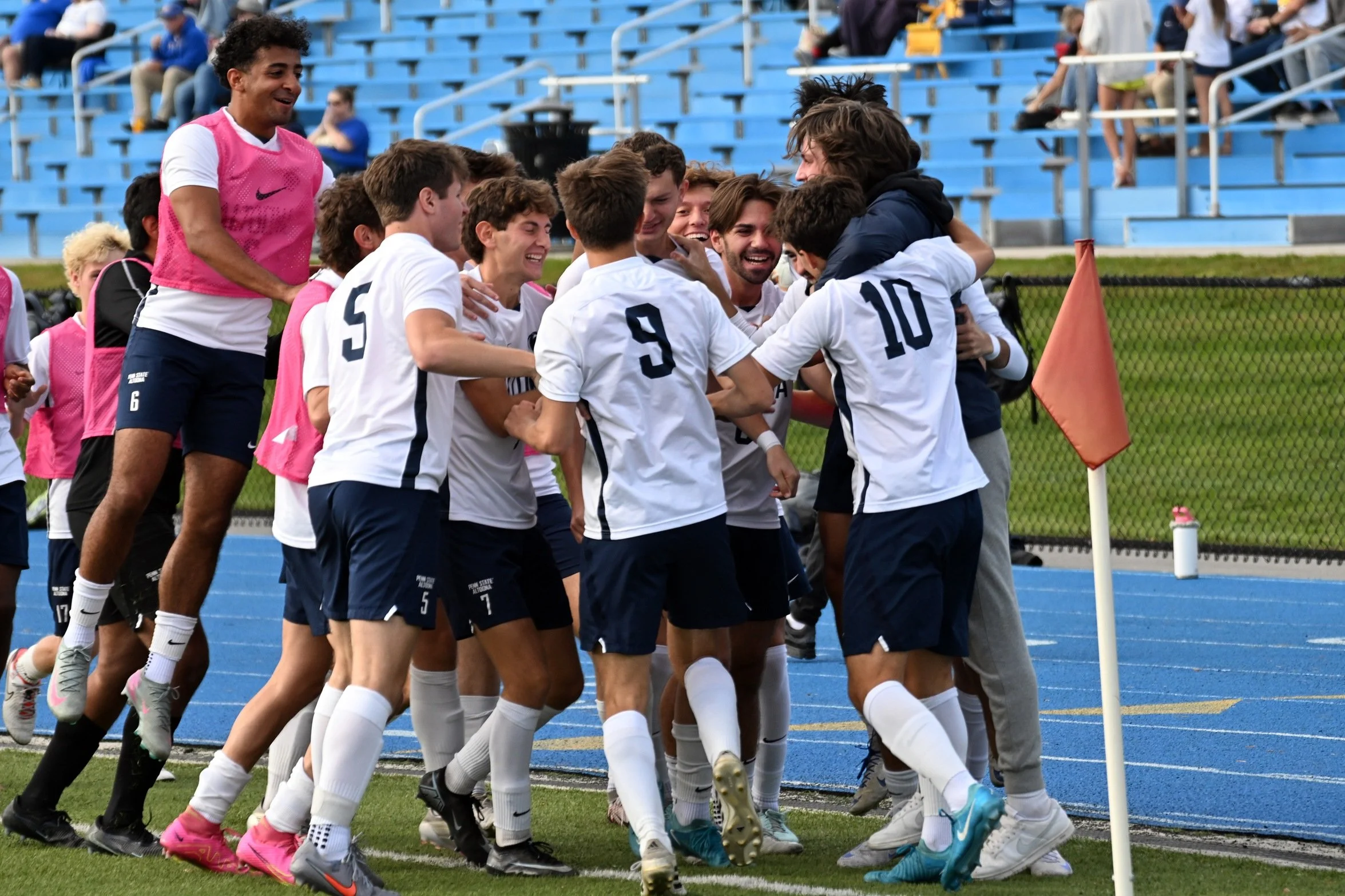 A group of soccer players celebrating and huddling together on the field near a corner flag, with players wearing white jerseys and others in pink bibs, in a stadium with blue seats in the background.