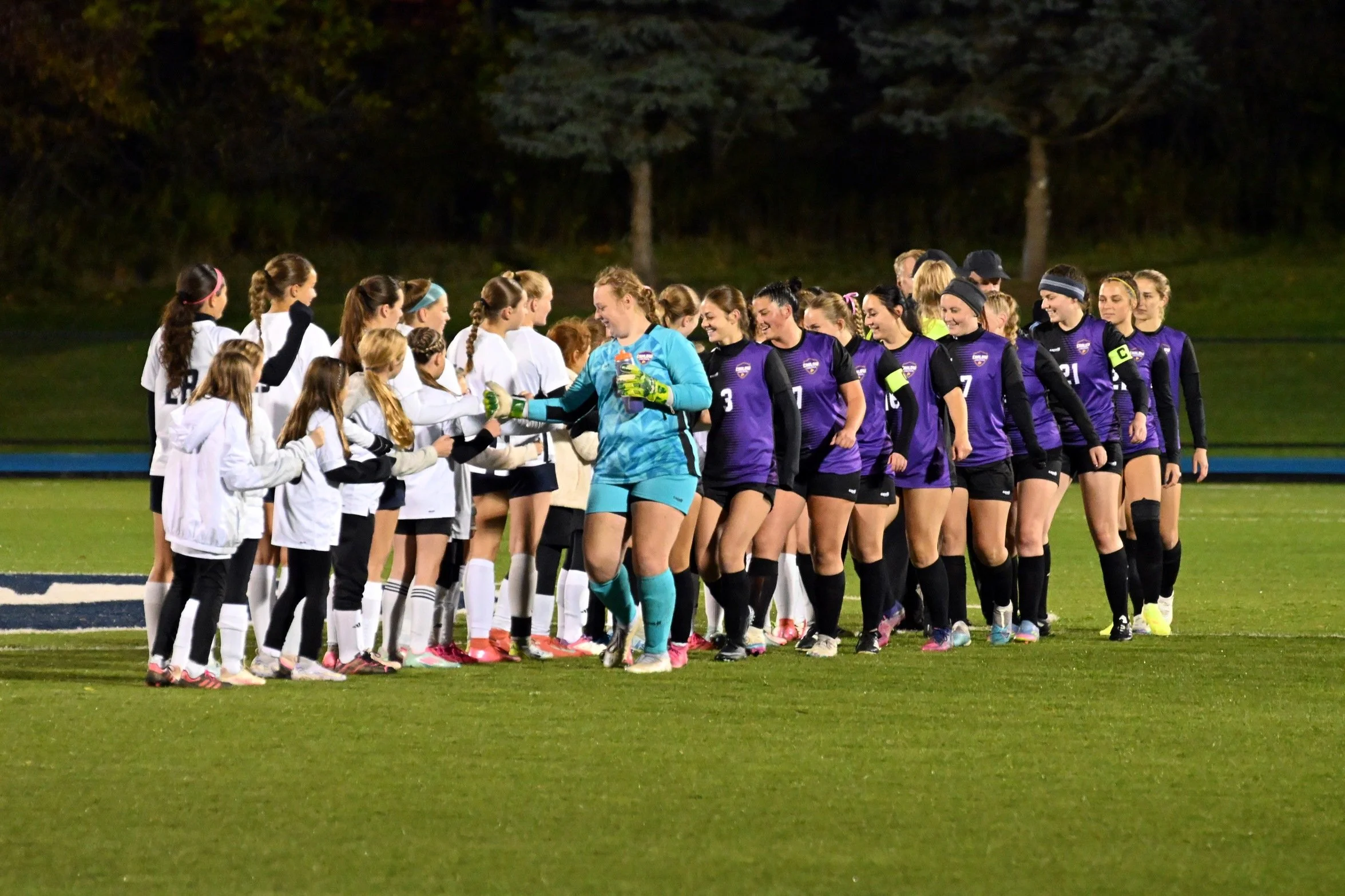 Women soccer players line up and shake hands on the field at night, with trees in the background.