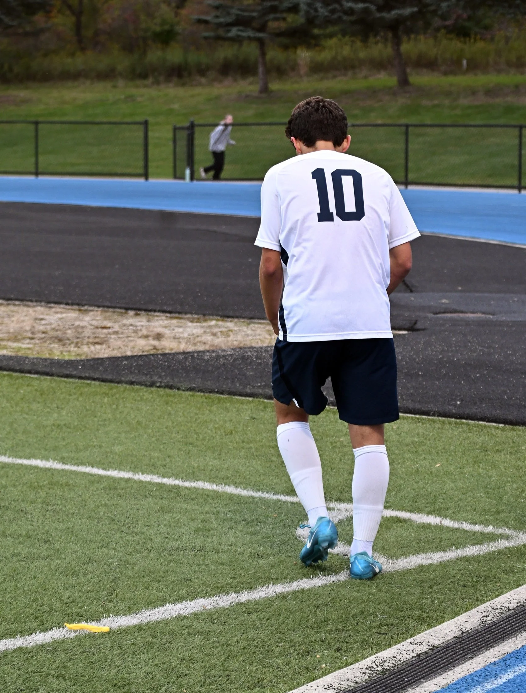 A soccer player wearing a white jersey with the number 10 on the back, blue cleats, and white socks, walking on the soccer field near the sideline while another person in a gray jacket runs in the background on the other side of the fence.