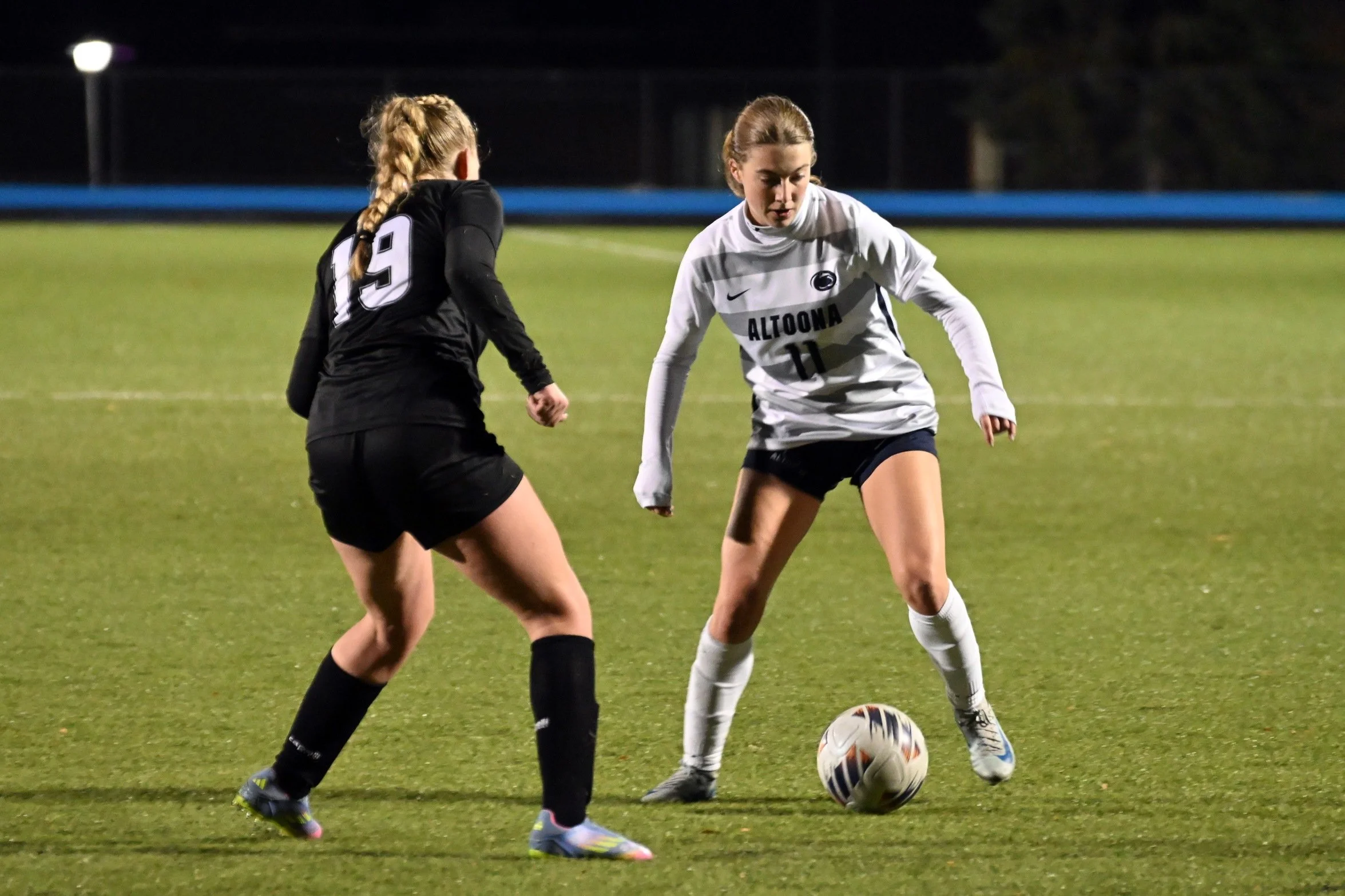 Two female soccer players compete for the ball on a grassy field during a night game, one wearing a black jersey with the number 19 and the other wearing a white jersey with the logo 'ALTOONA' and the number 11.